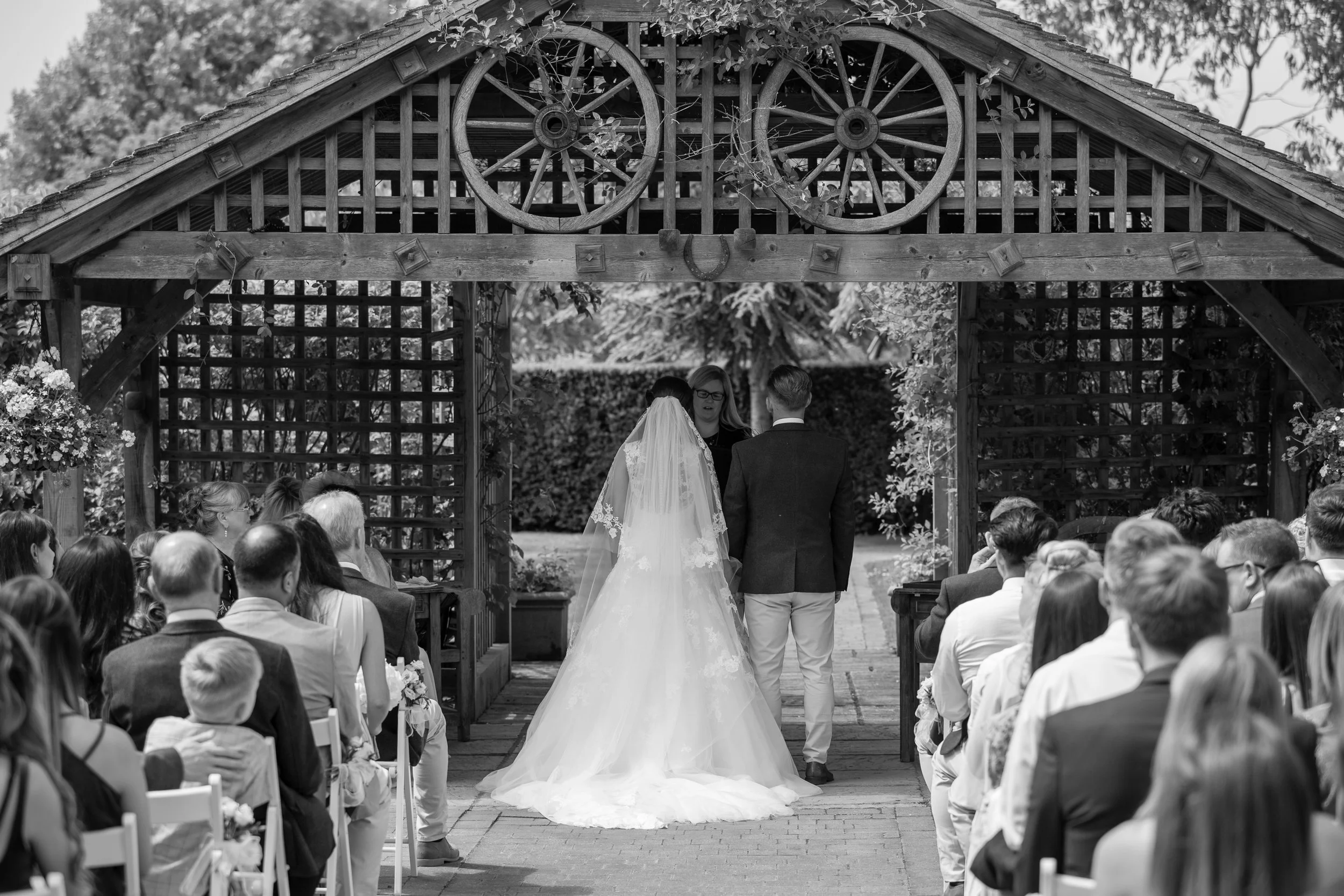 A black and white photo of a wedding ceremony taking place outdoors under a wooden arch with wagon wheels at the top. The bride and groom stand facing each other at the altar, with an officiant in between. Guests are seated on both sides, watching the ceremony in a garden setting with trees and bushes in the background.