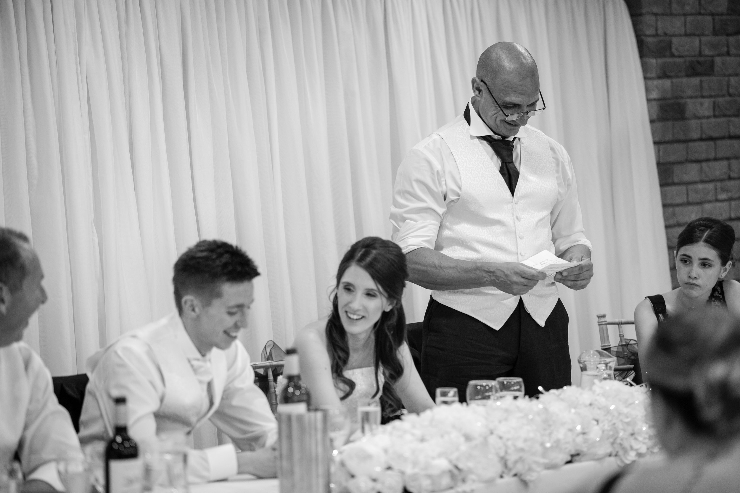 A man giving a speech at a wedding reception table with smiling guests, black and white photo.