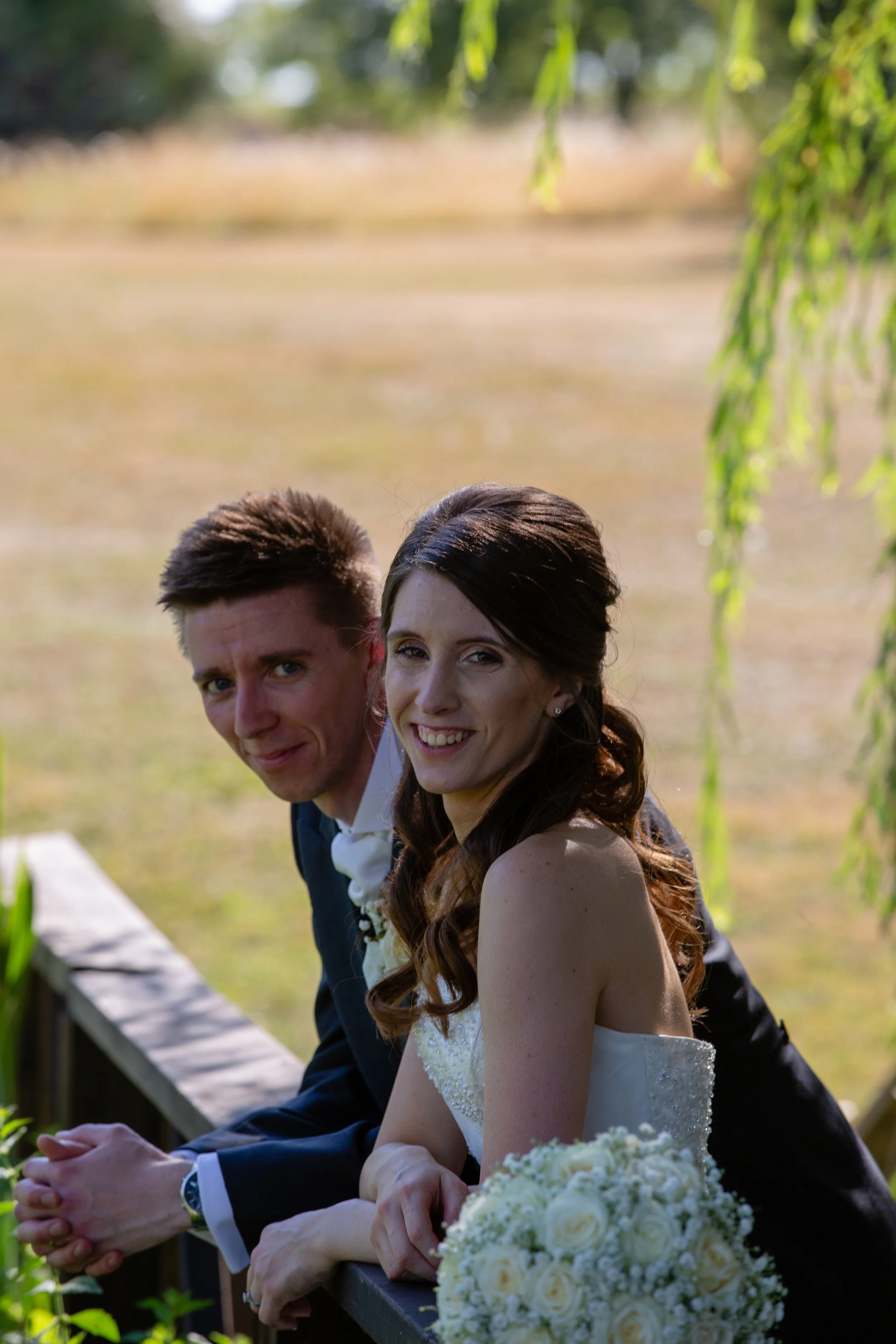 A newlywed couple on a wooden bridge outdoors, smiling and holding hands, with a blurred natural background.