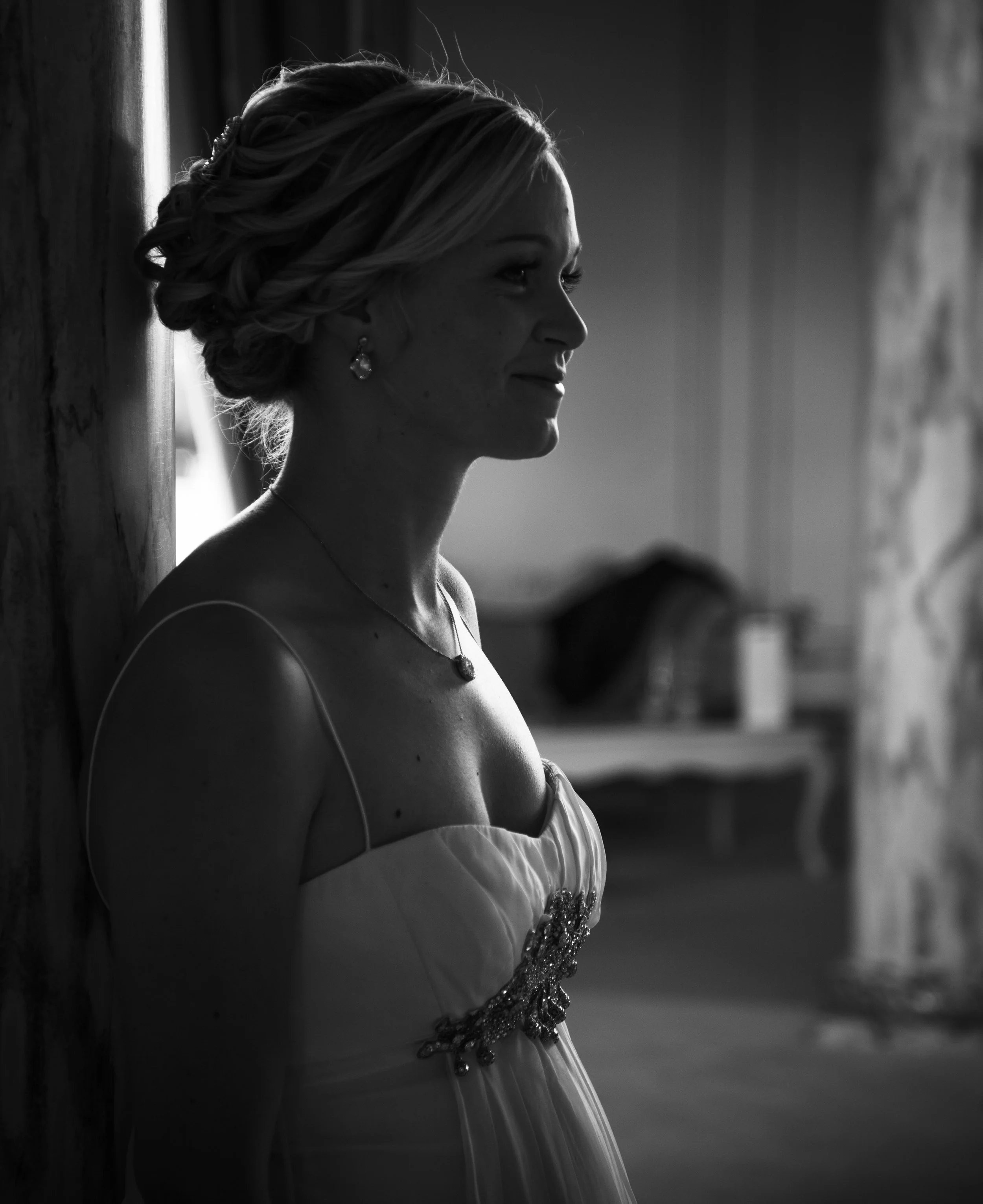 Profile of a woman in a strapless dress with jewelry, leaning against a wall, in a softly lit room.