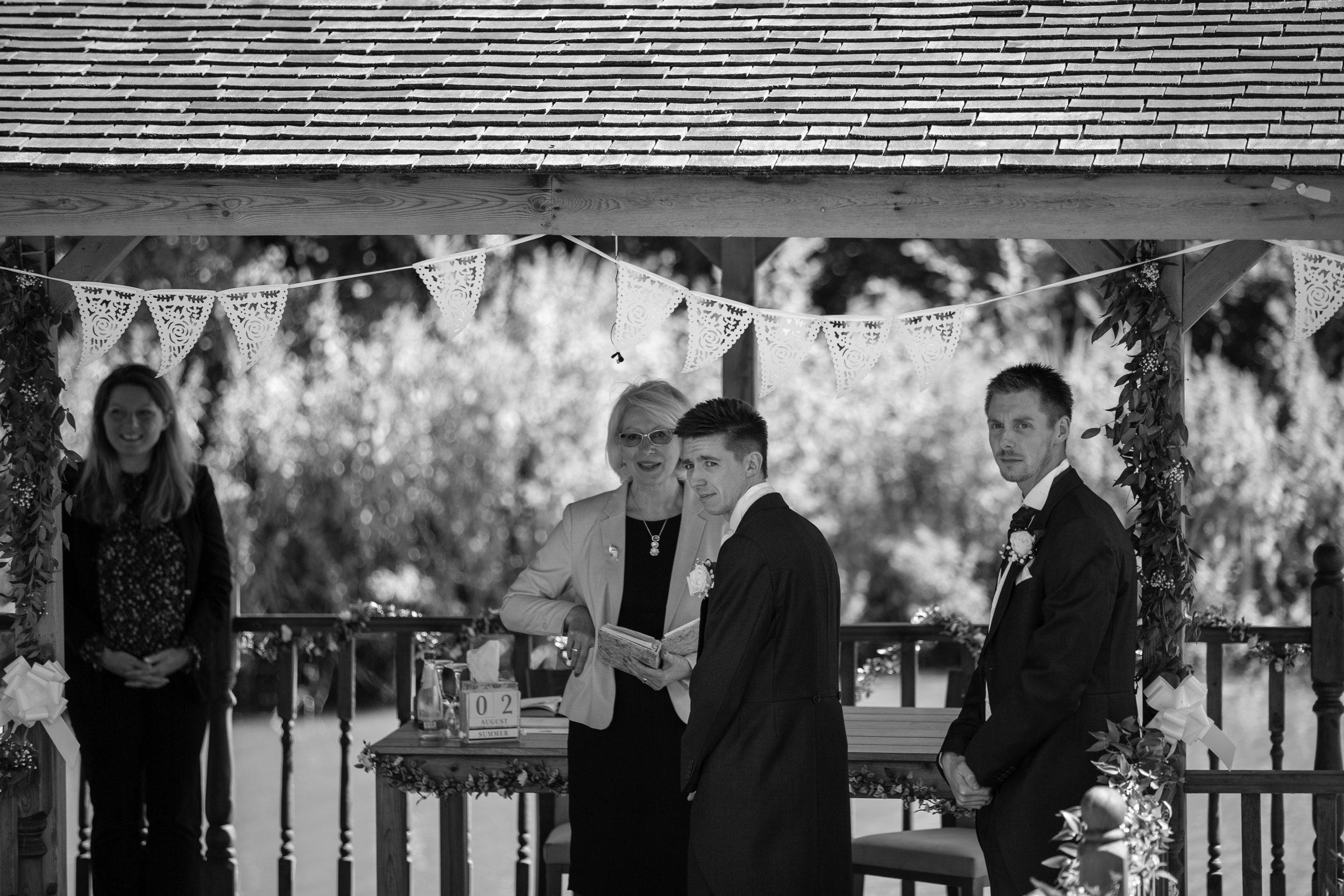 A black and white photo of a wedding ceremony outdoors. The bride and groom are standing under a decorated wooden arch, with a woman officiant reading from a book. Two other people, a woman on the left and a man on the right, are standing nearby, all