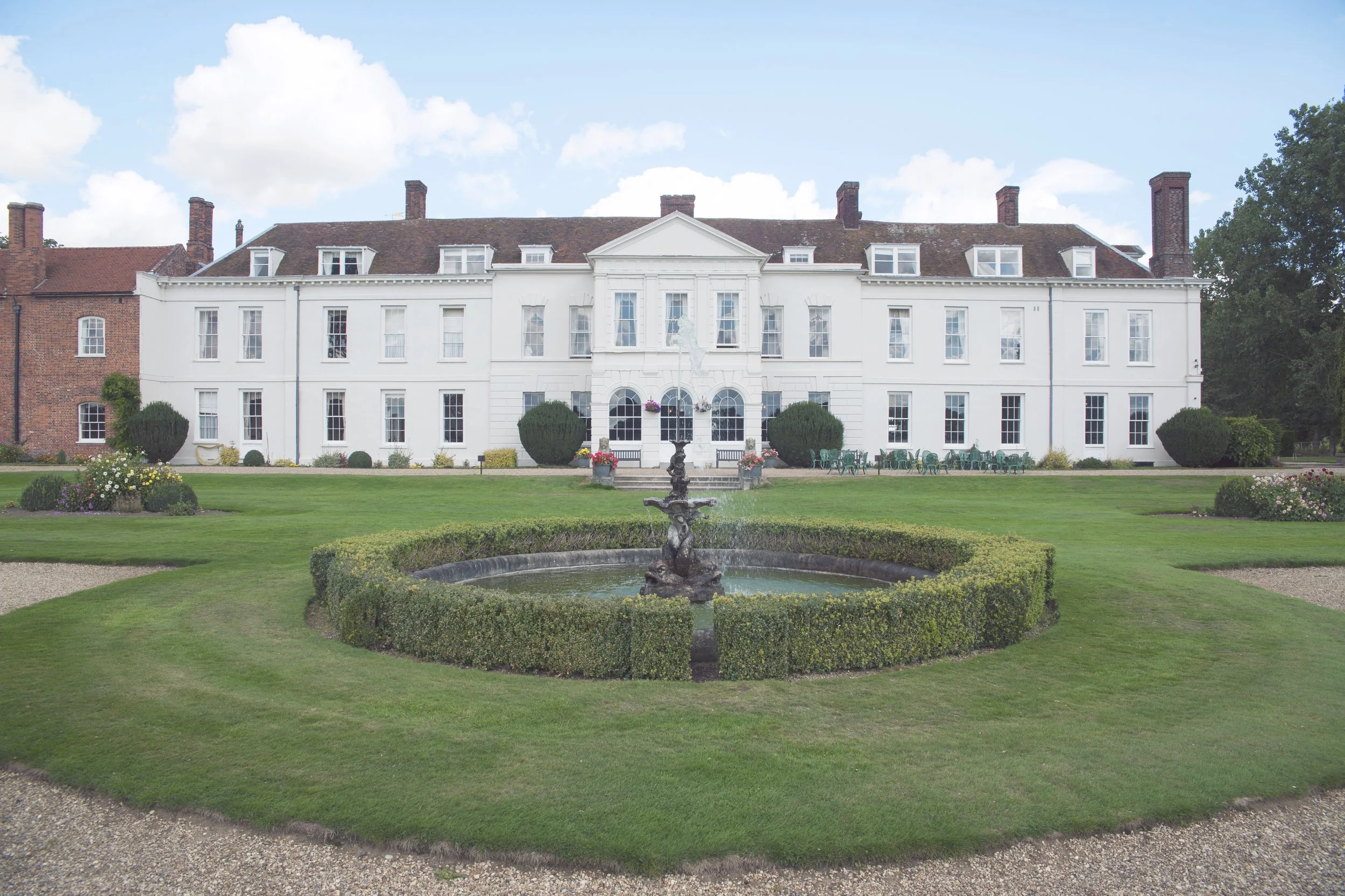 A large white historic mansion with multiple windows and chimneys, surrounded by a landscaped garden with a fountain in the foreground, green grass, bushes, and trees under a partly cloudy sky.