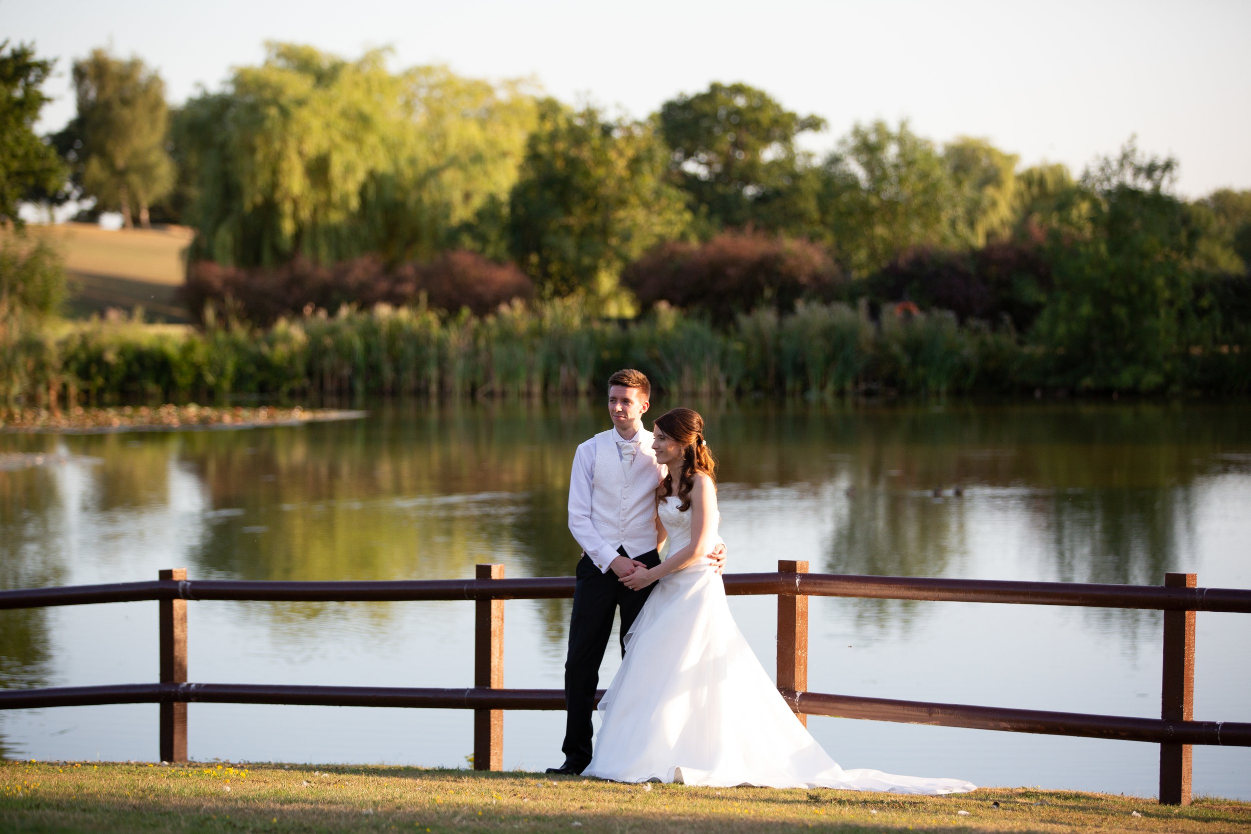 a newlywed couple standing by a lake at sunset, with trees in the background