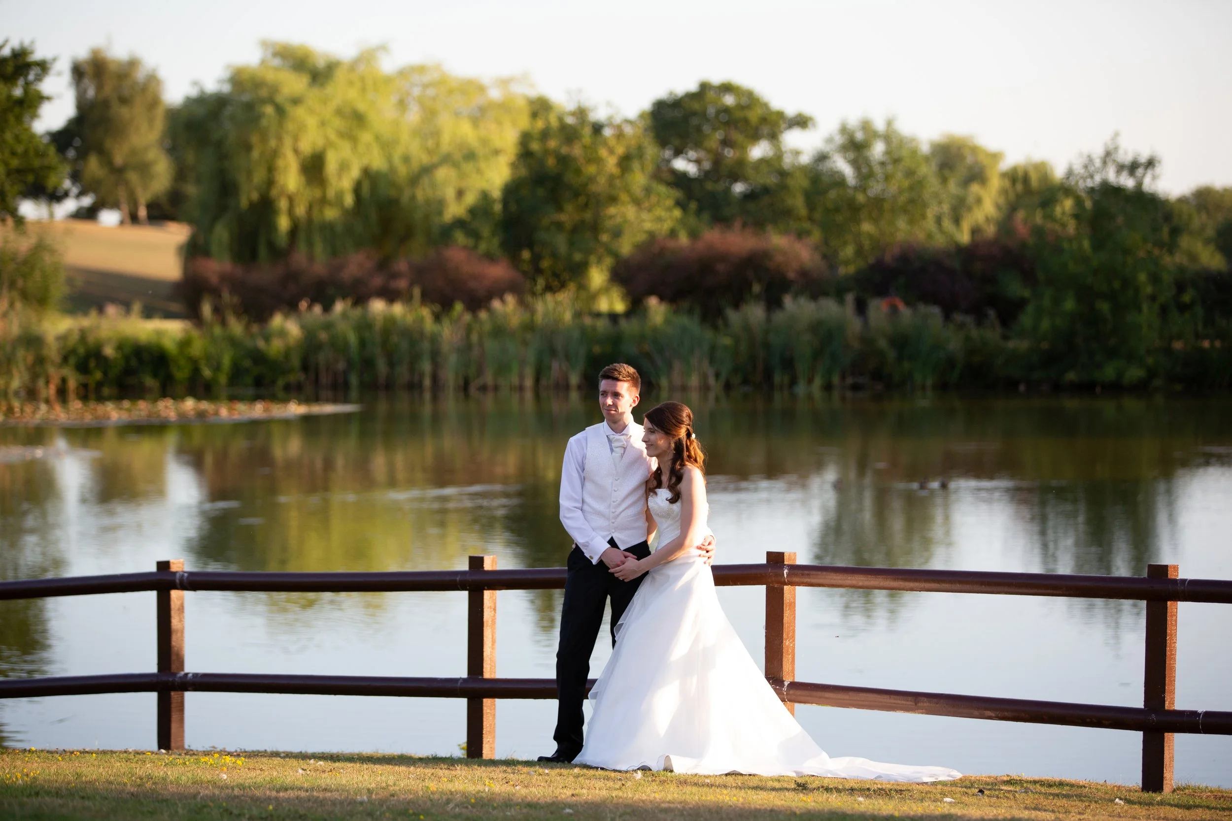 a newlywed couple standing by a lake at sunset, with trees in the background