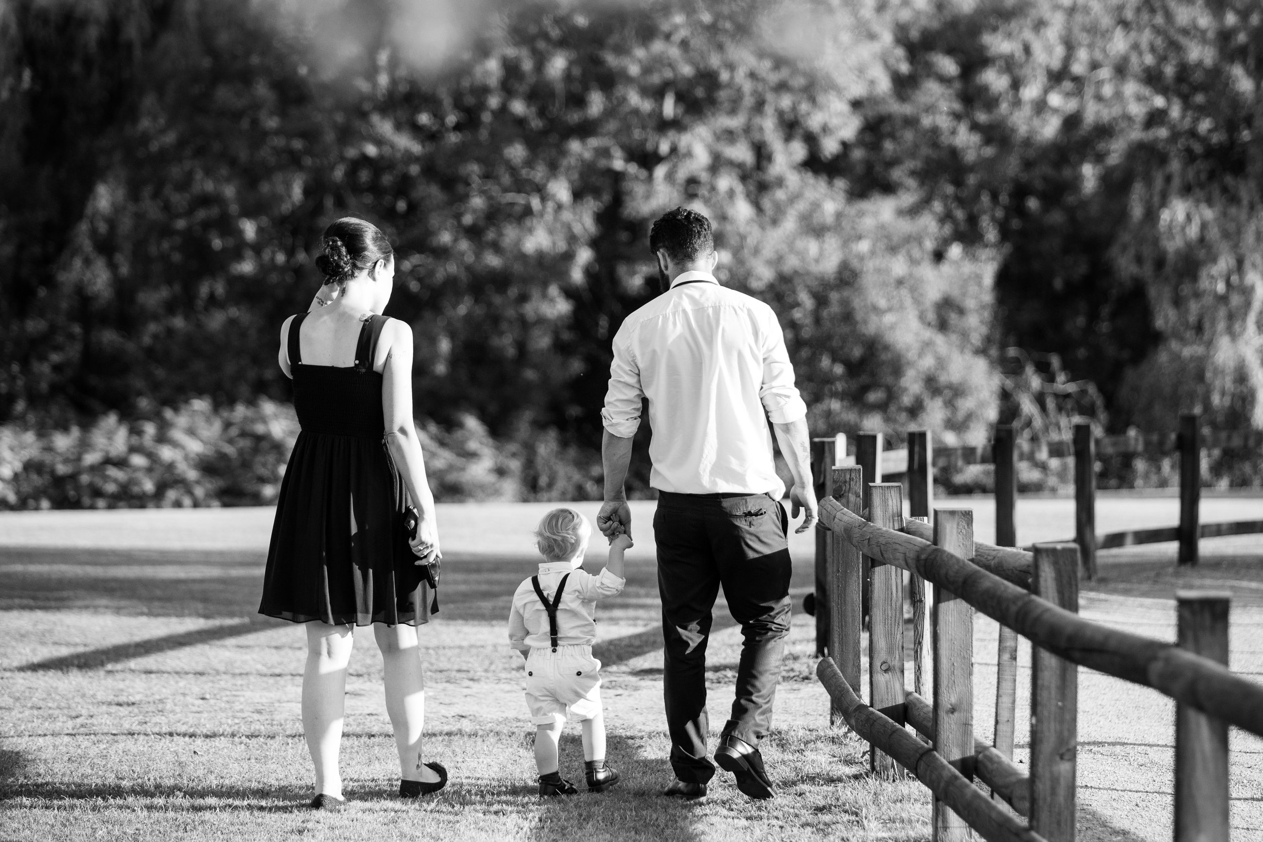 A family of three walking hand in hand outdoors in a park, with a woman in a dress, a man in a shirt, and a young child dressed in white shorts and suspenders.