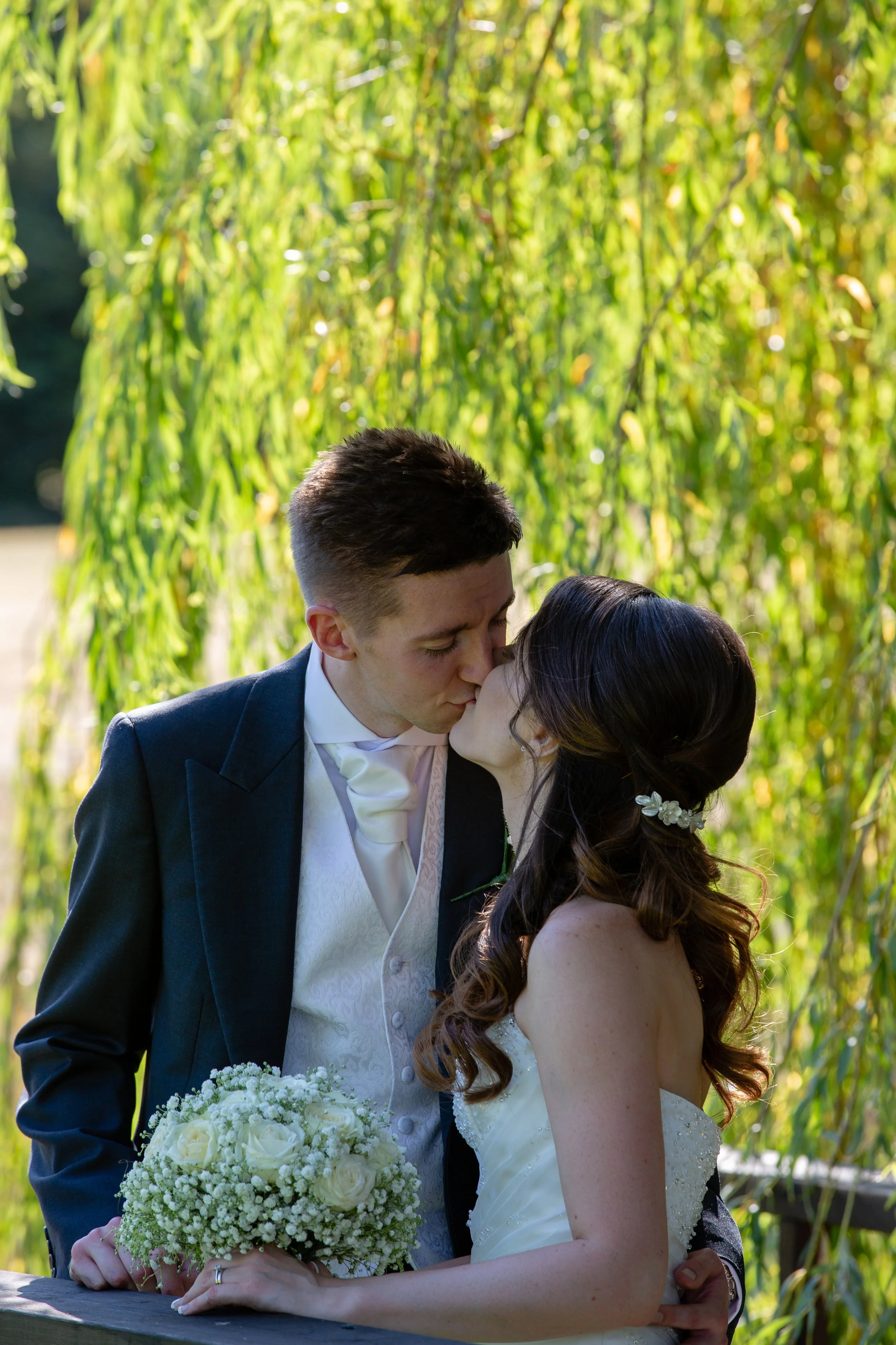 A bride and groom sharing a kiss outdoors, with green foliage in the background. The bride holds a bouquet of white flowers. The groom wears a dark suit and vest, while the bride has long wavy hair with a floral hair accessory.