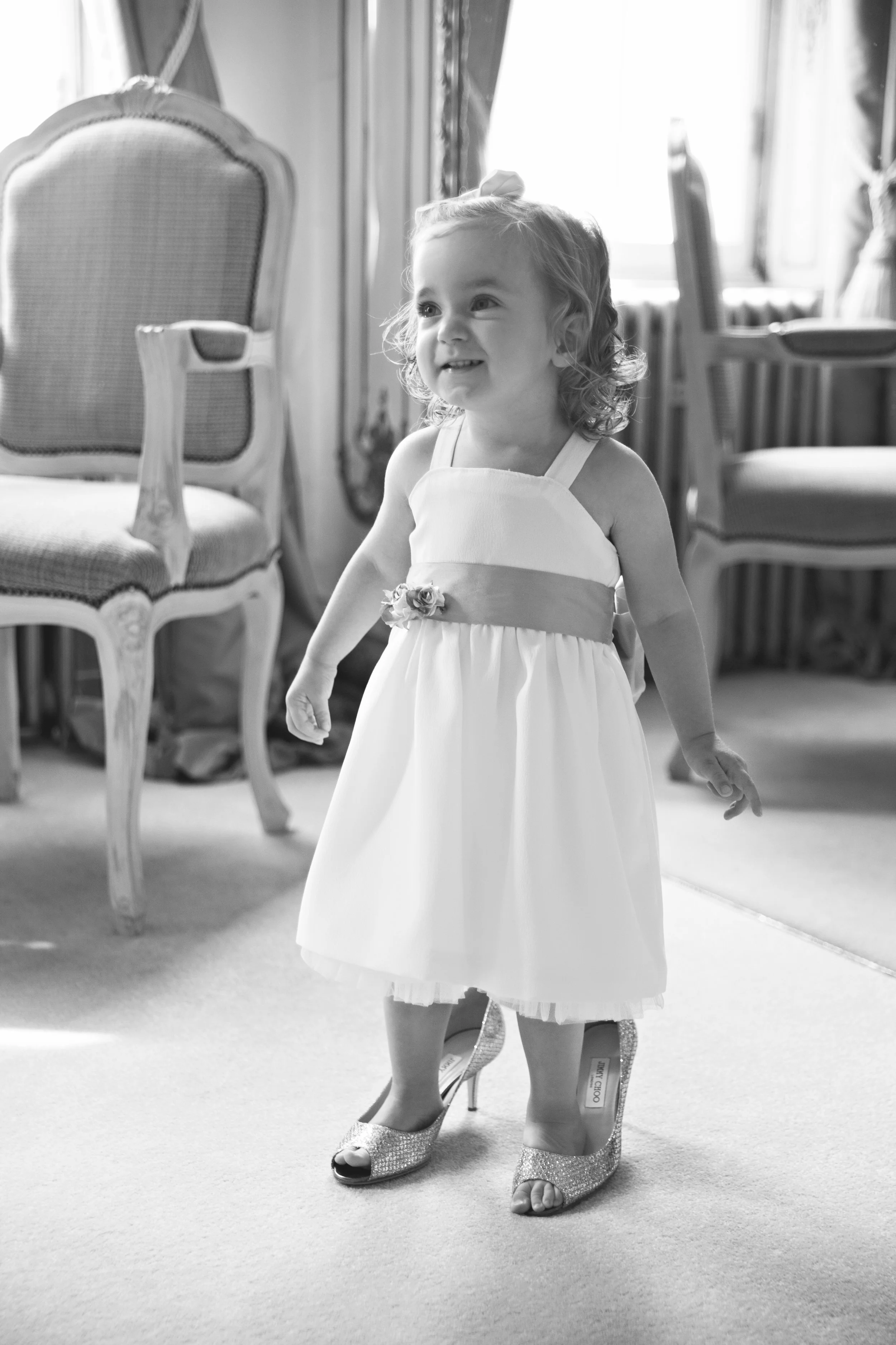 Black and white photo of a young girl in a dress and high heels standing indoors, smiling and looking to the side, with chairs and curtains in the background.