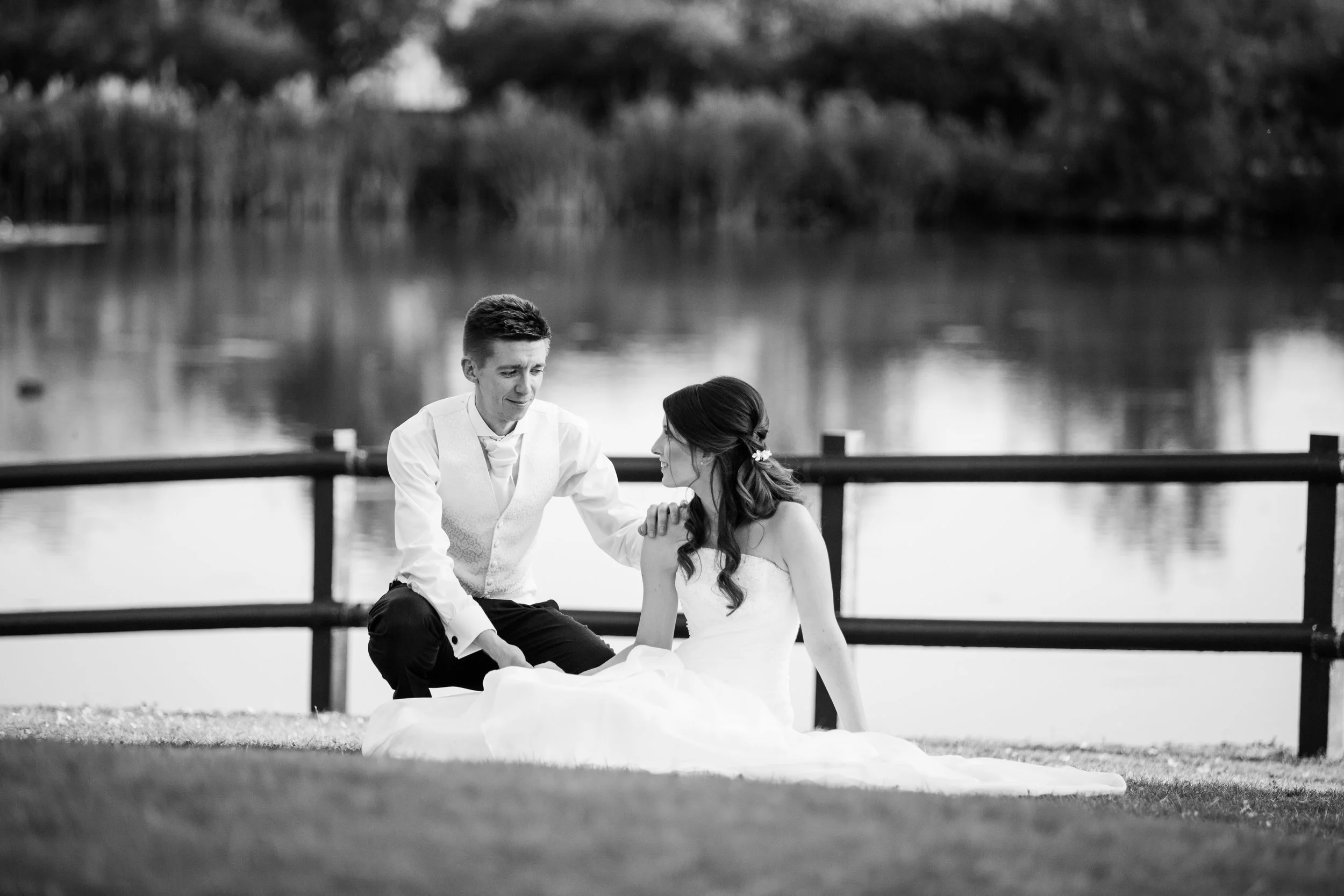 A black-and-white photo of a bride and groom by a lakeside. The bride, sitting on the grass, is wearing a strapless wedding dress with long dark hair styled in loose waves, holding the groom's hand. The groom, kneeling next to her, is dressed in a wh