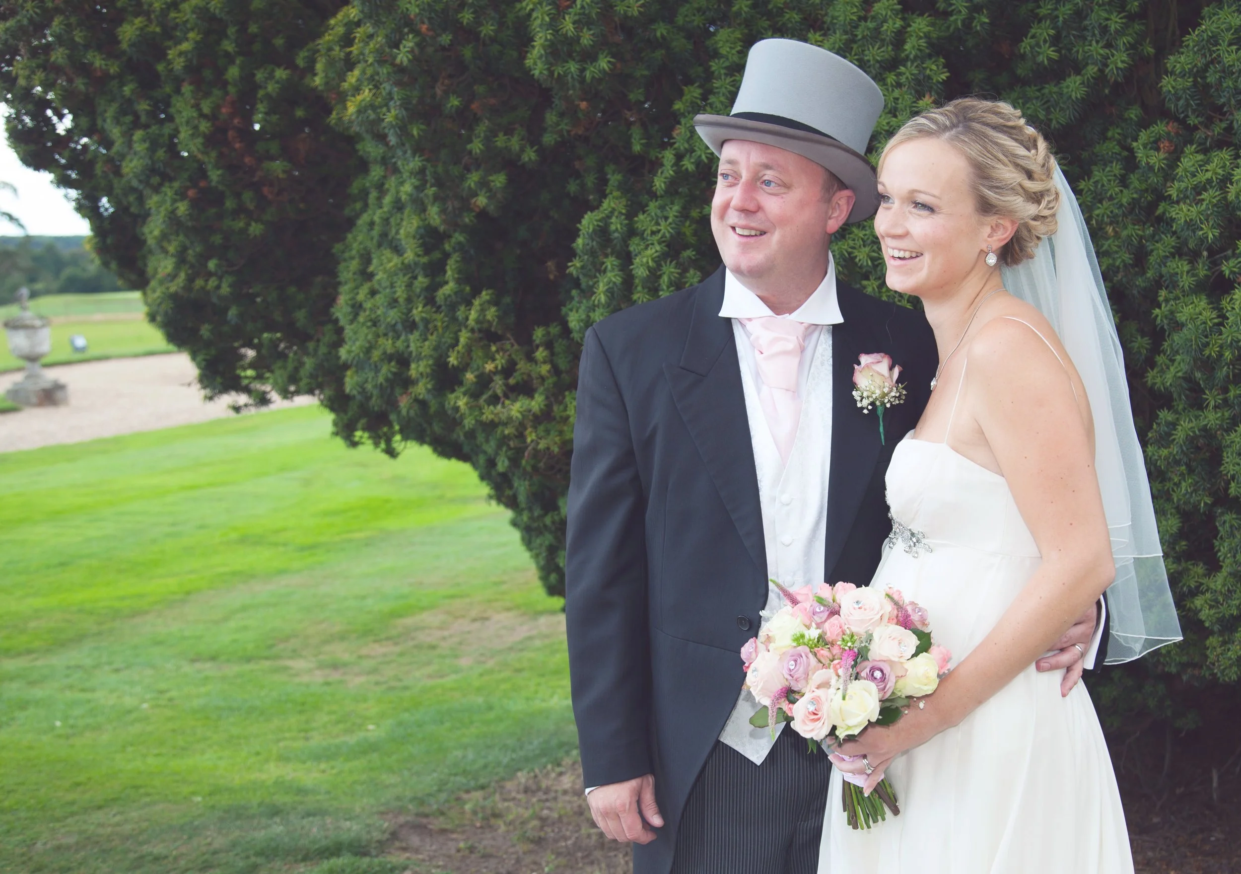 A bride and groom on their wedding day stand outdoors near a large green bush. The groom wears a black suit with a top hat, and the bride wears a white wedding dress and holds a bouquet of pink and white roses. They are smiling and looking to the sid
