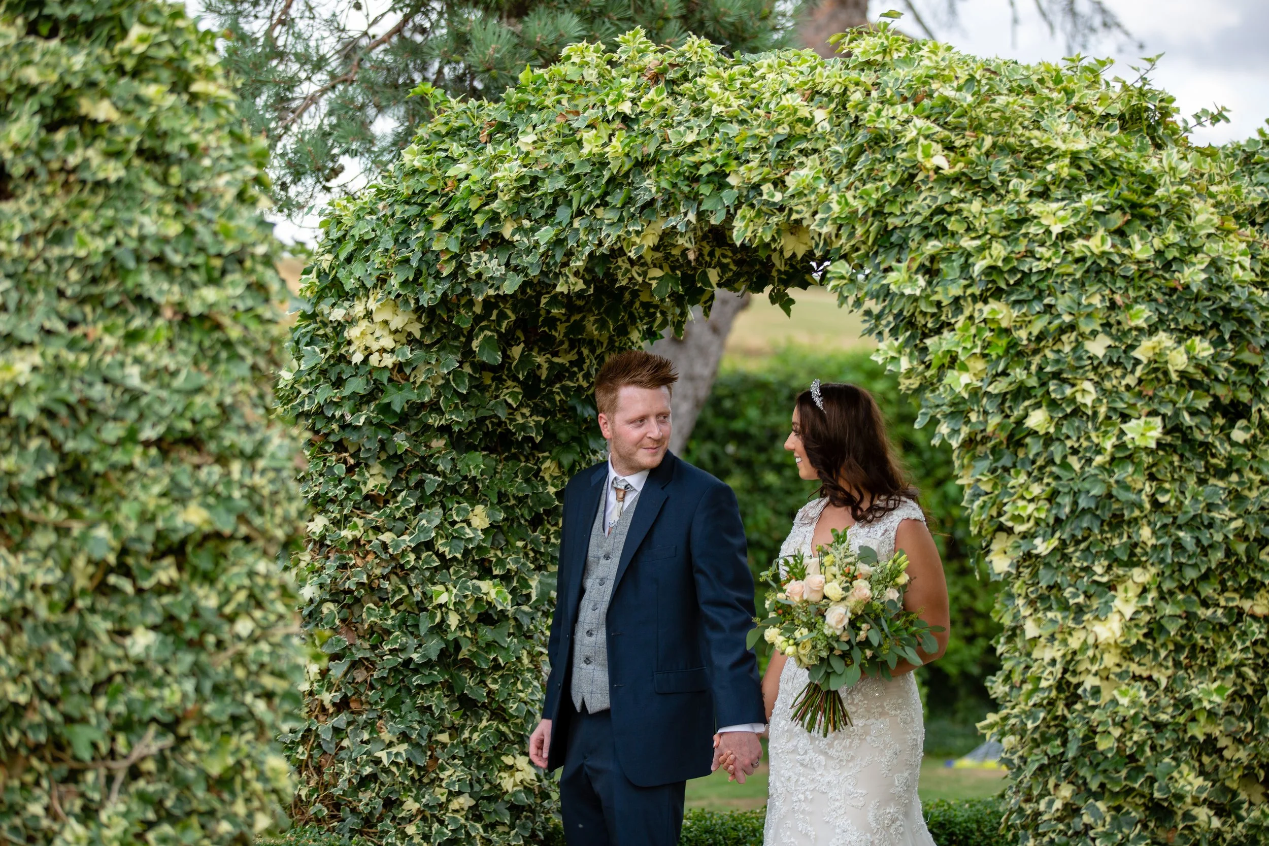 A newlywed couple holding hands and looking at each other, standing under a green, leafy arch during their outdoor wedding.