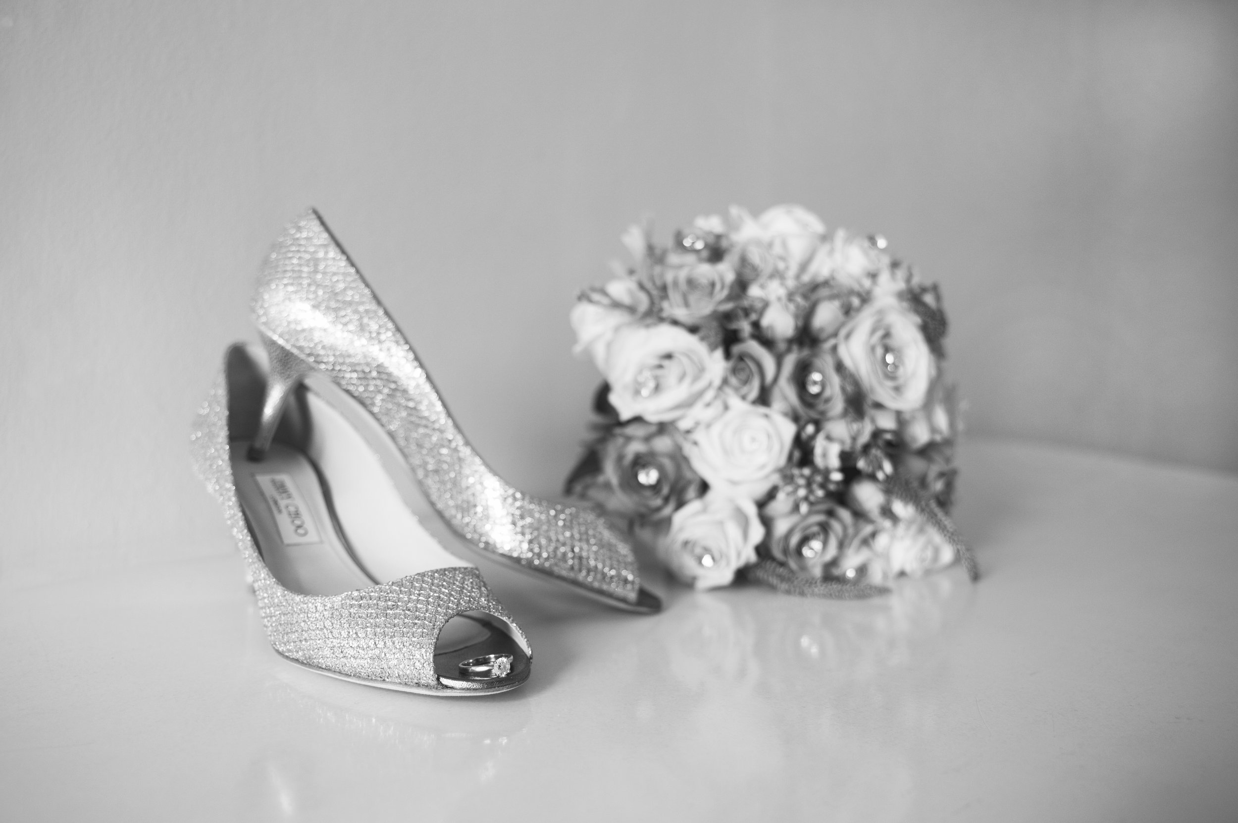 A pair of sparkly high heels, a wedding band inside the open toe of one shoe, and a bouquet of roses and decorative accents, all in black and white.