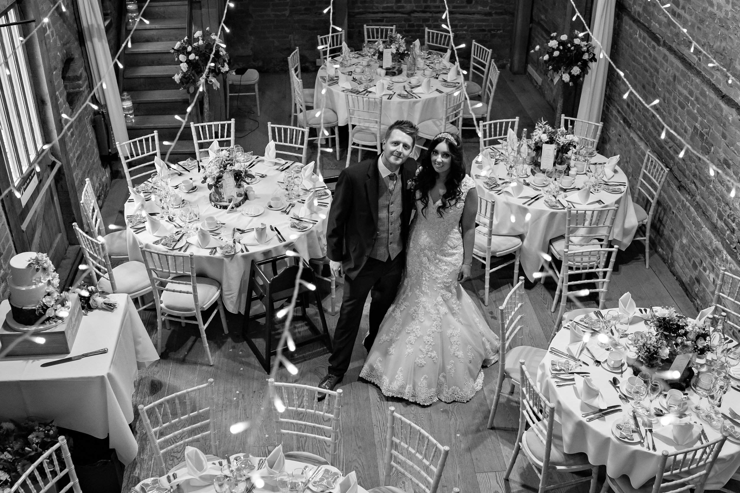 A black-and-white photo of a bride and groom standing together in a decorated wedding reception hall. The hall has round tables set with tableware and floral centerpieces, chairs, and string lights hanging above. The bride is wearing a detailed lace 