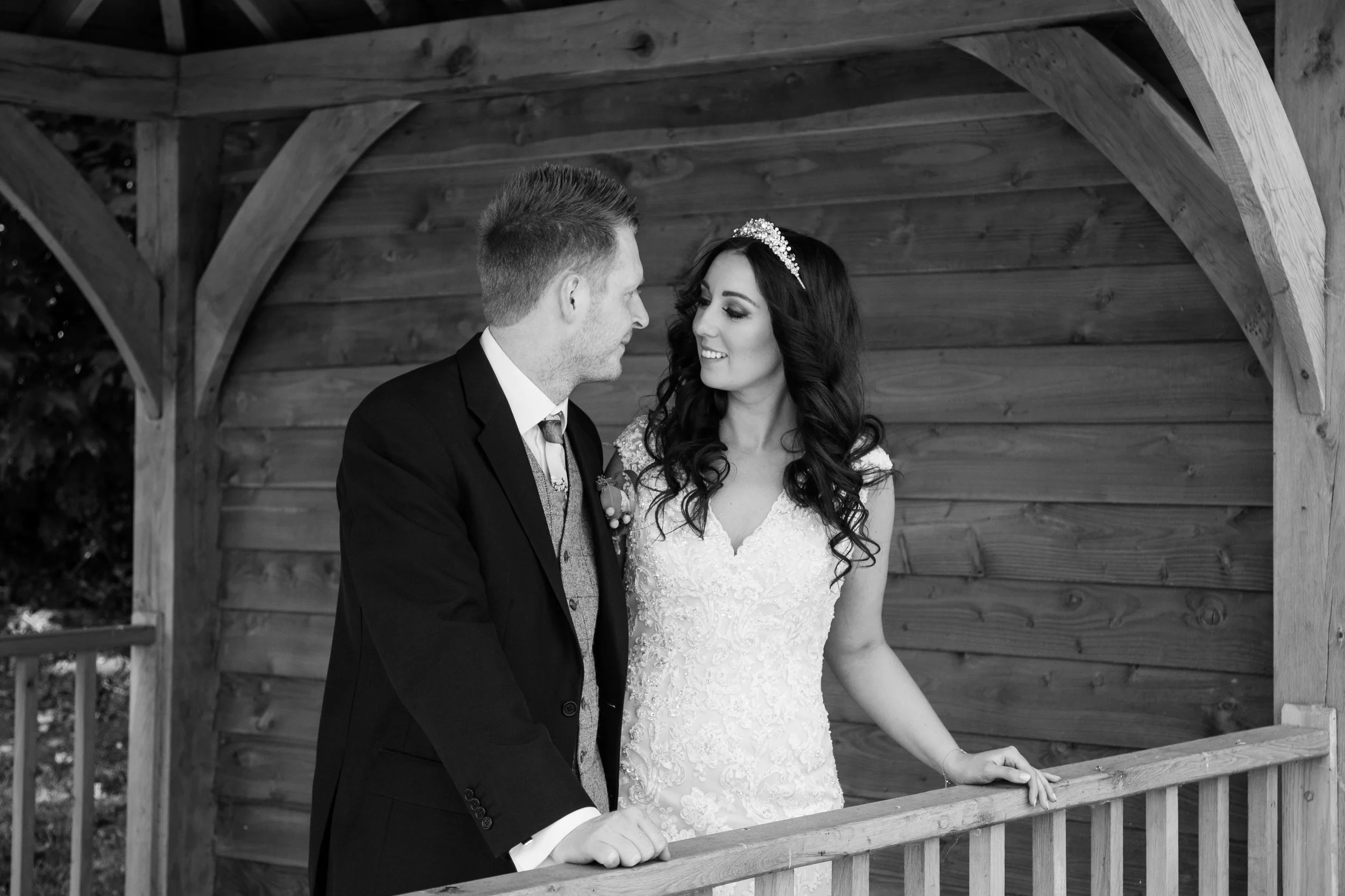 A black and white photo of a bride and groom standing on a wooden balcony, looking at each other affectionately.