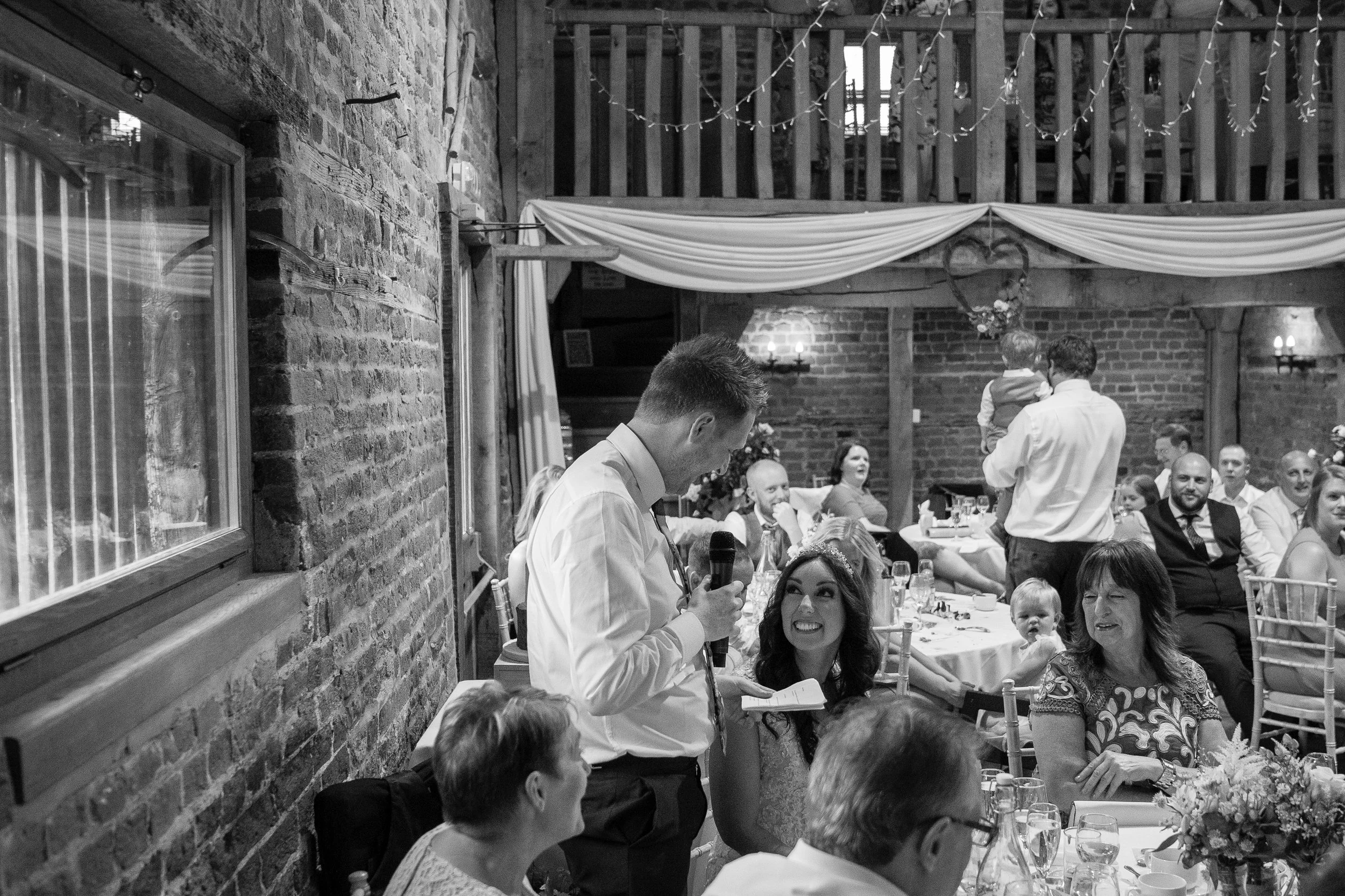 A wedding reception with people sitting at tables, listening to a man giving a speech with a microphone, a woman smiling at him, and guests enjoying the event in a rustic venue decorated with drapes and string lights.