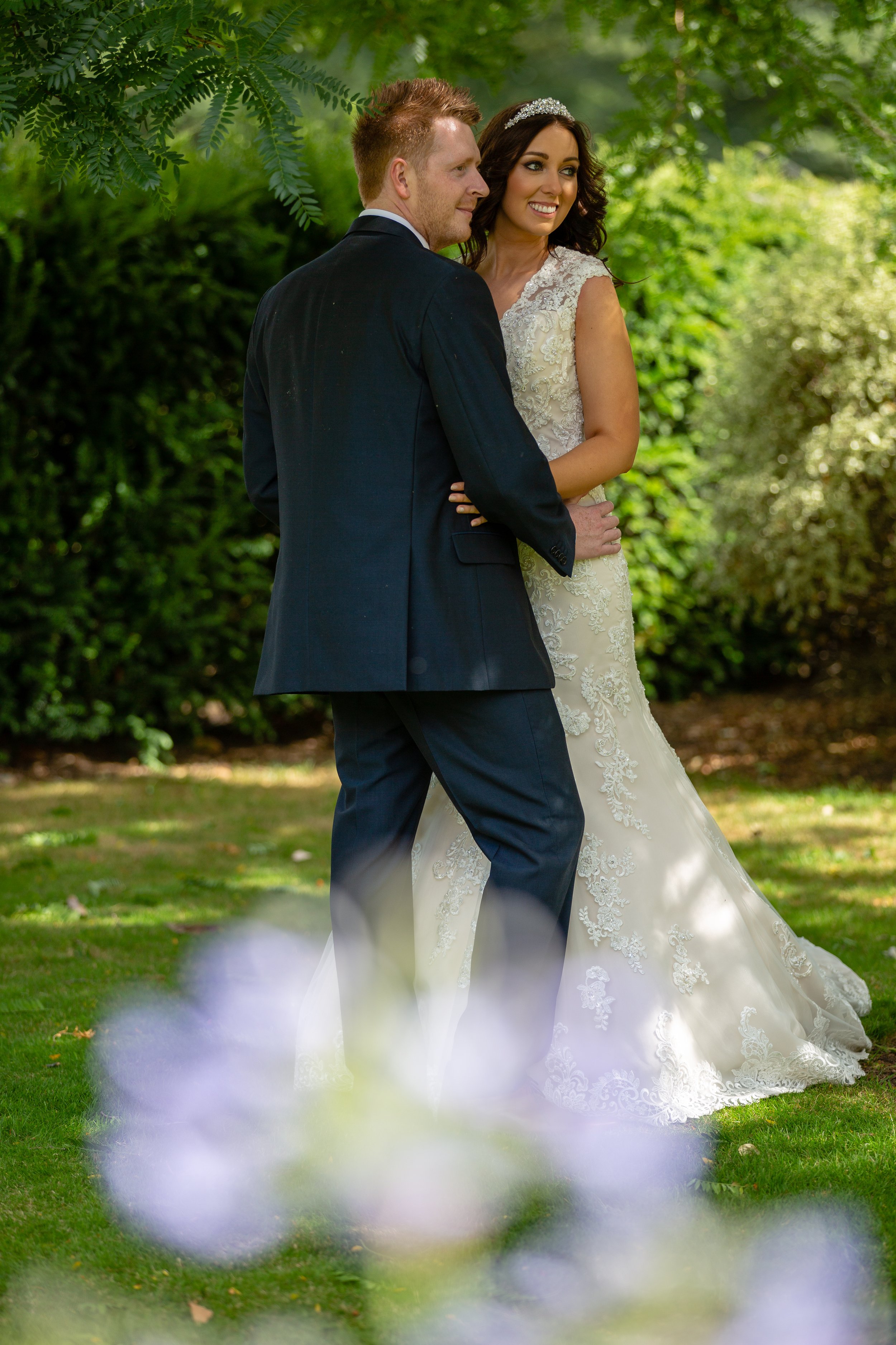 A bride and groom pose outdoors in a lush green garden, with the bride wearing a lace wedding gown and the groom in a navy suit.