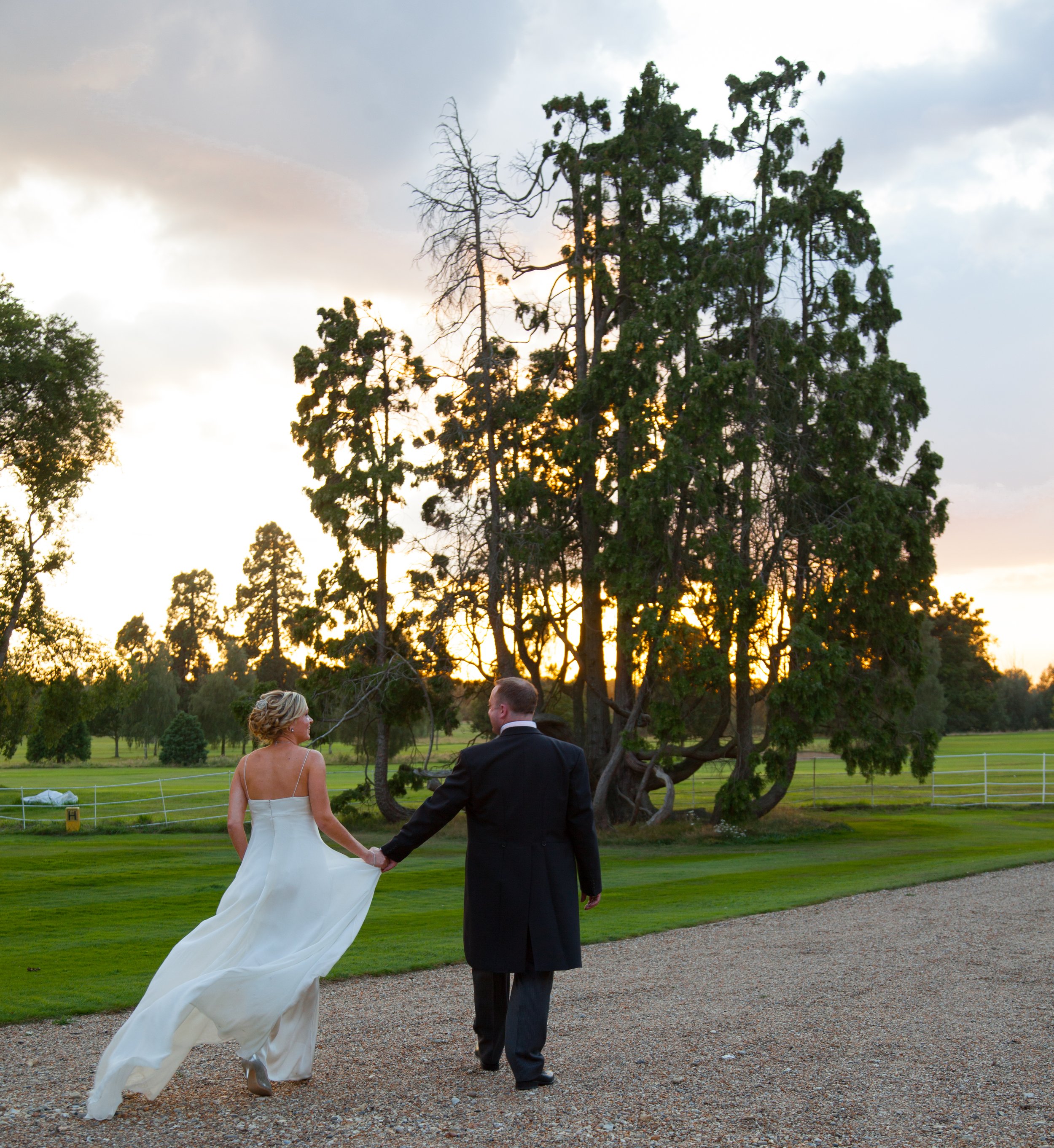 A bride and groom walking hand in hand on a gravel path in a park, with large trees and a sunset sky in the background.