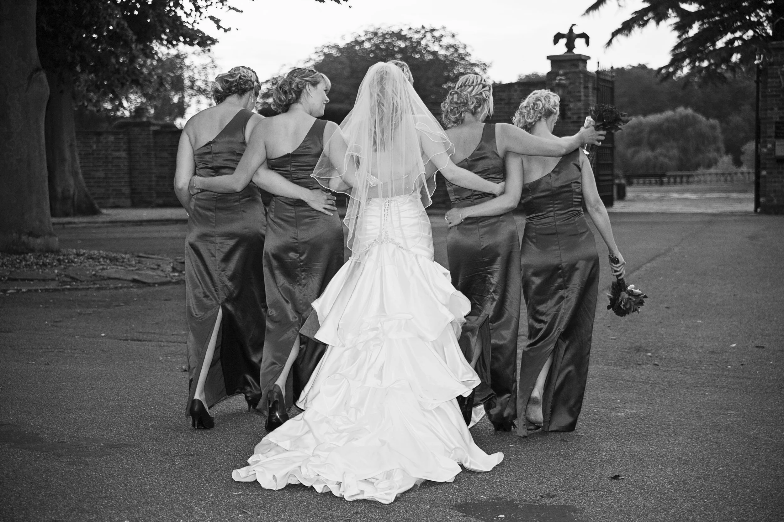 Black and white photo of a bride and bridesmaids walking outdoors, seen from behind. The bride wears a white wedding gown and veil, while the bridesmaids wear matching dark satin dresses, some holding bouquets. They are near a brick wall and trees.