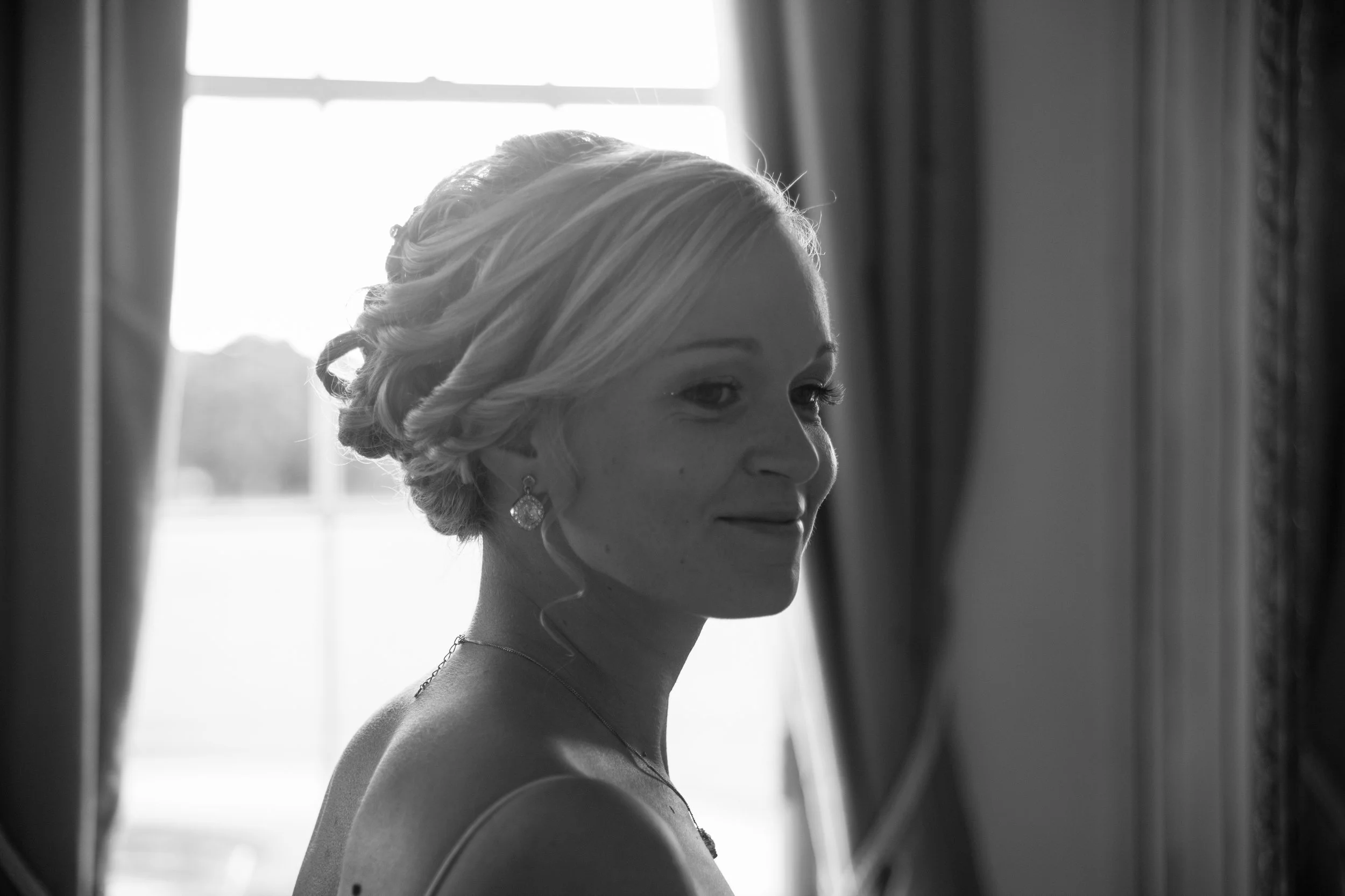 Black and white photo of a smiling woman with styled hair, earrings, and a necklace, standing near a window, indoors.