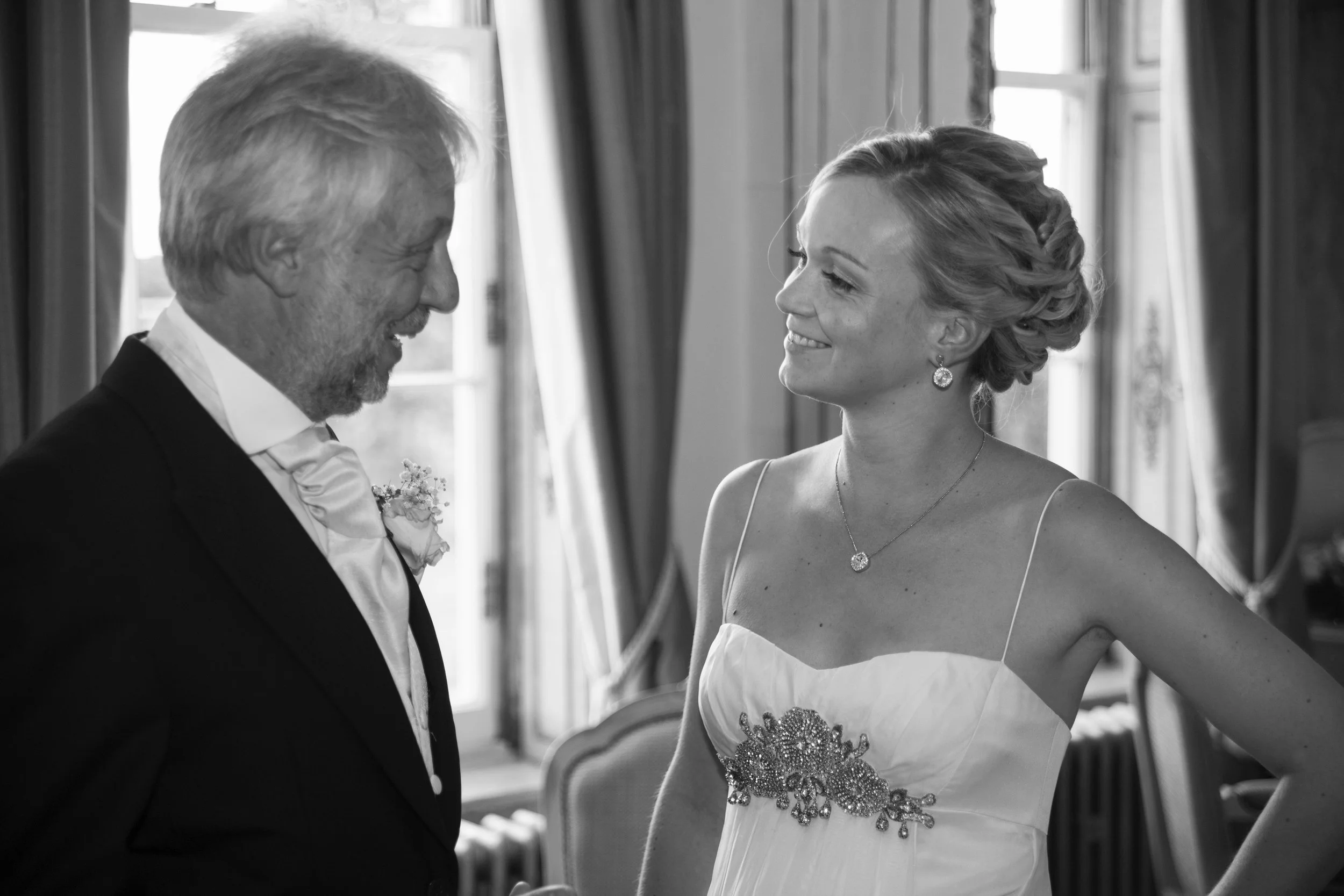 A black and white photo of an older man and a young woman smiling at each other, indoors near windows with curtains, likely at a wedding or special event.