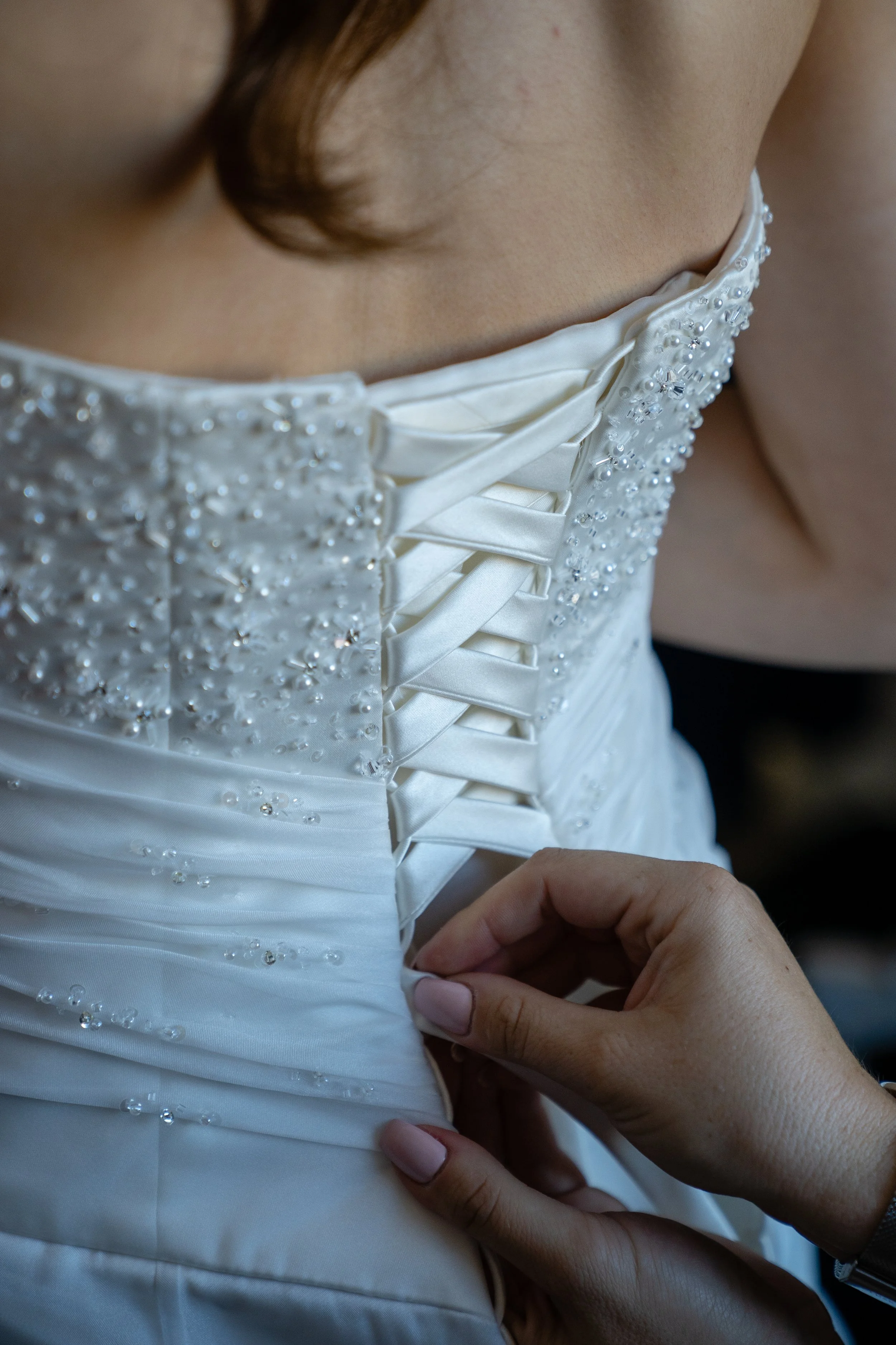 Close-up of a woman fastening the laces on her white wedding gown, which is decorated with pearls and beads.