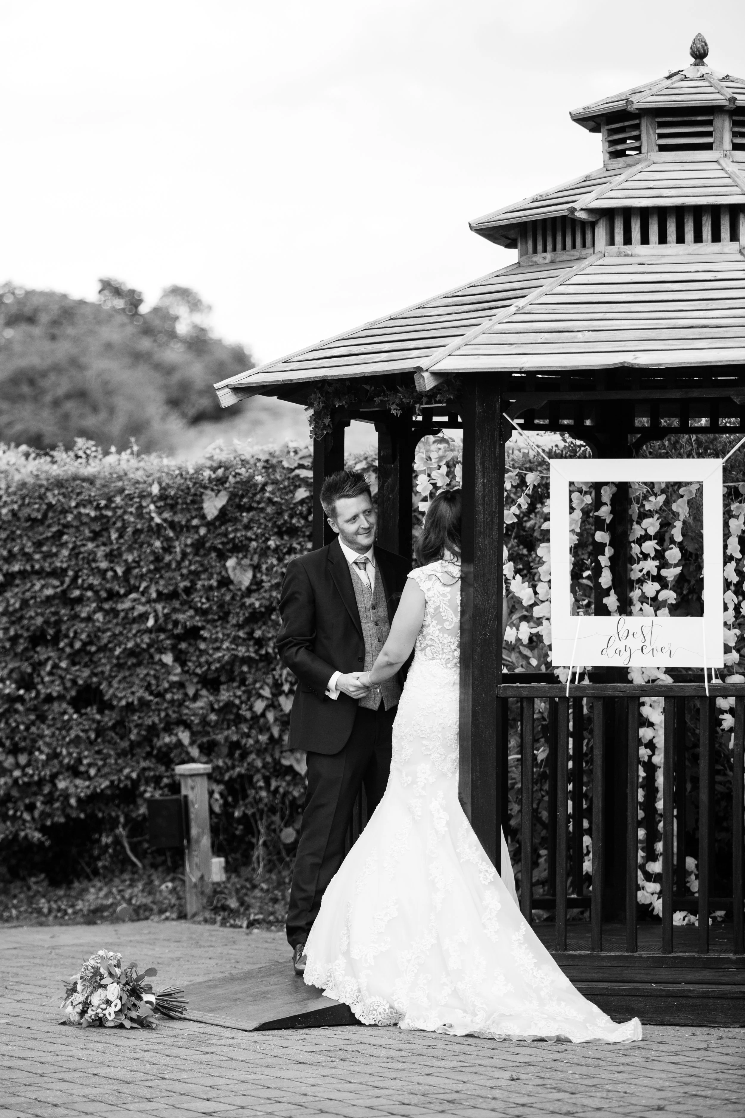 A black and white photo of a bride and groom holding hands during their wedding ceremony outdoors near a small gazebo. The bride is wearing a lace wedding dress, and the groom is dressed in a suit. A bouquet is on the ground nearby, and a sign on the