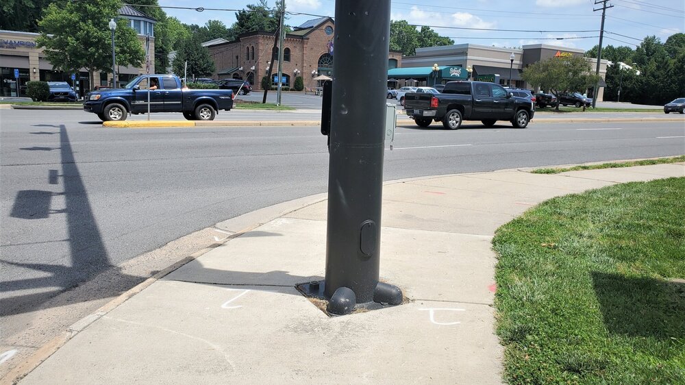A traffic light pole with a “beg button” for crossing is placed in the middle of the sidewalk.