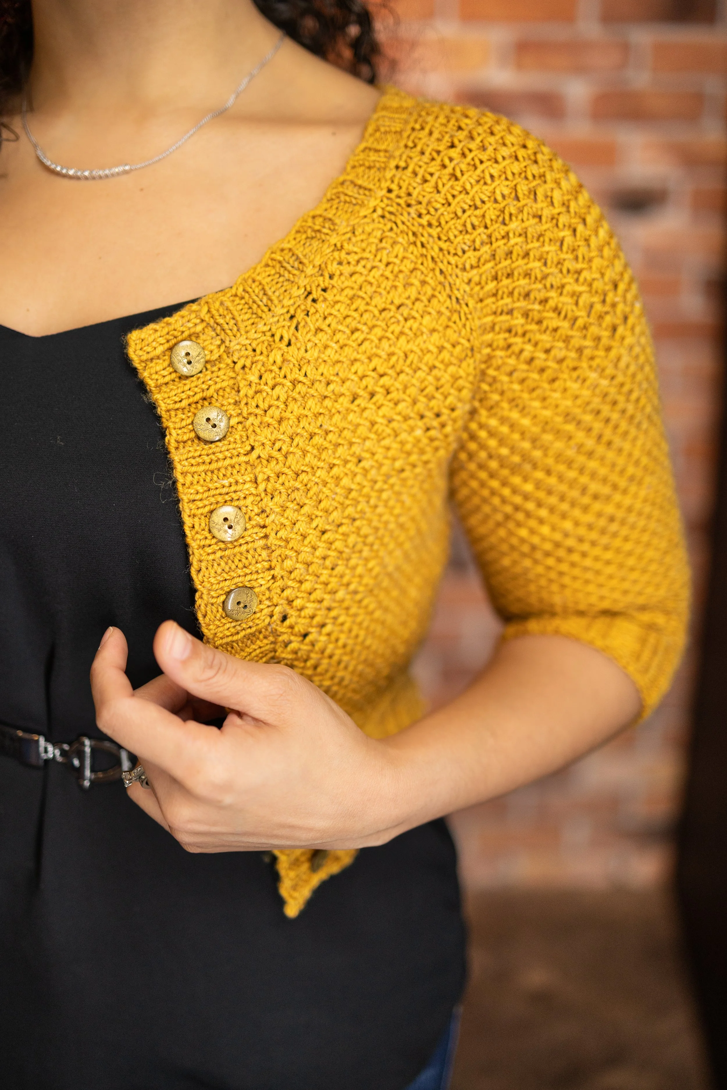A closeup of the squishy texture and button band on a cropped, little yellow sweater, showing off its cute buttons and neat finishing. Nydia Cardigan PDF knitting pattern by Vanessa Smith Designs.