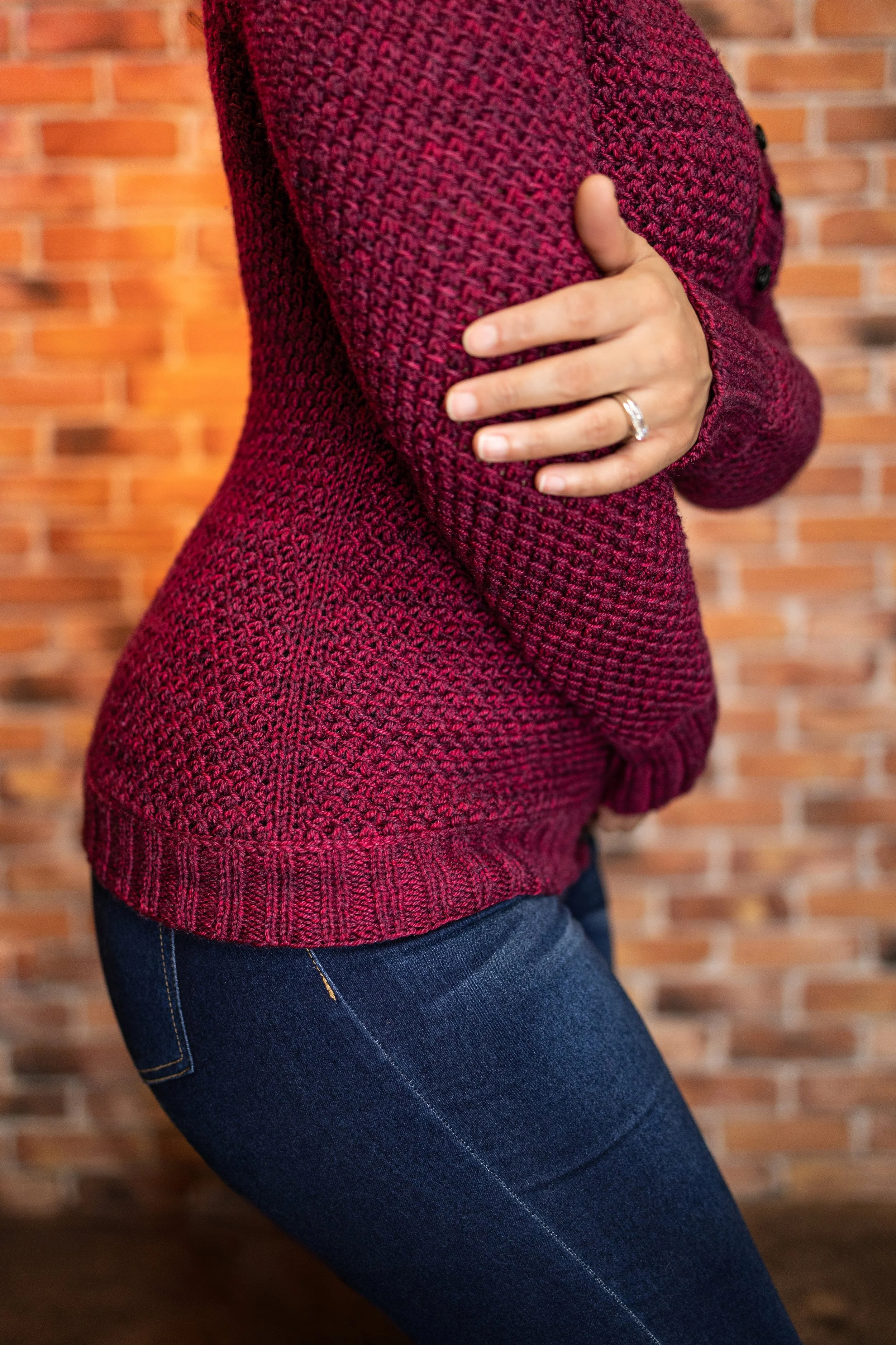 Side profile view of the full-length red Nydia Cardigan by Vanessa Smith, showing its close fit and intricate details: an easy allover textured lace stitch pattern, Vikkel/Estonian braids, and ribbing