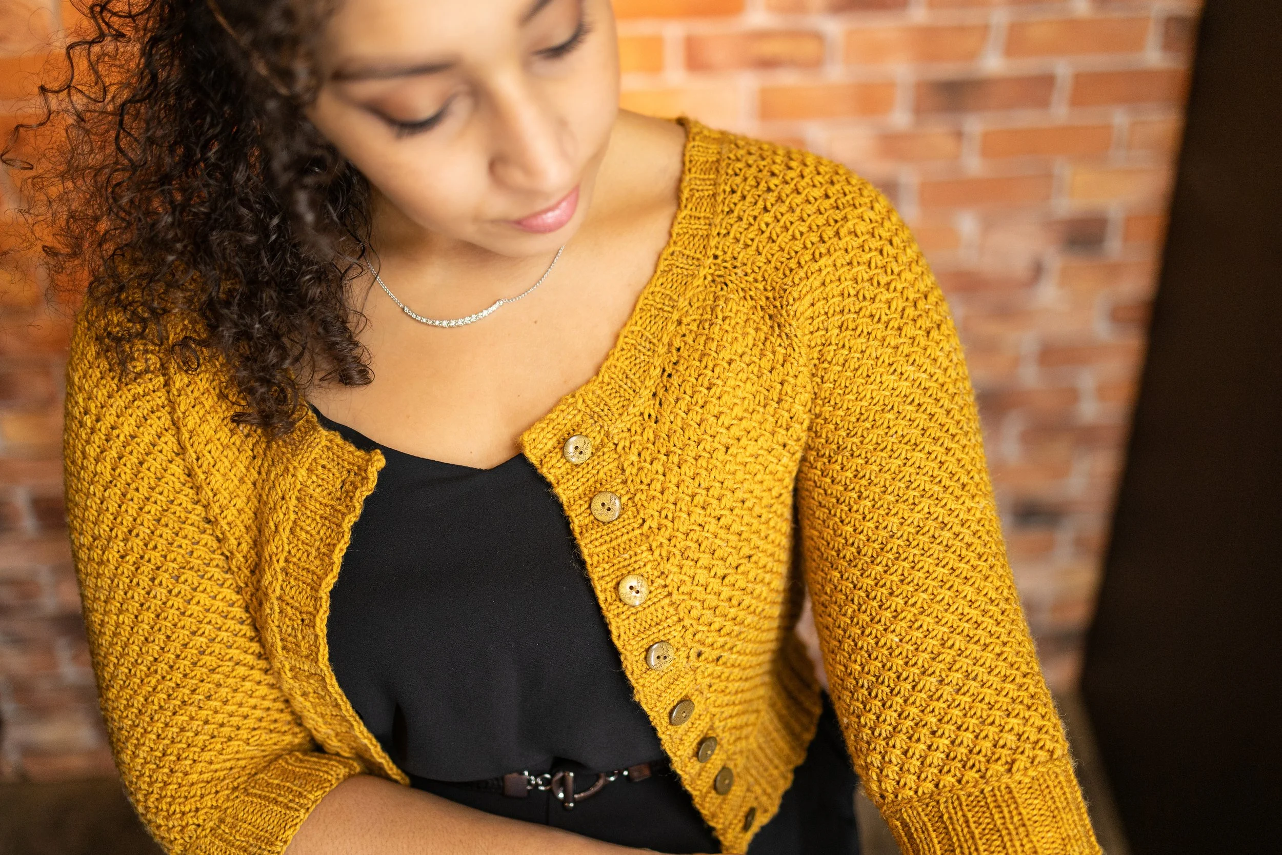 A curly-haired, biracial woman shows the details of the beautiful all-over textured lace stitch pattern and classic 2x2 ribbing on the cropped Nydia Cardigan. PDF knitting pattern by NessaReneeKnits.