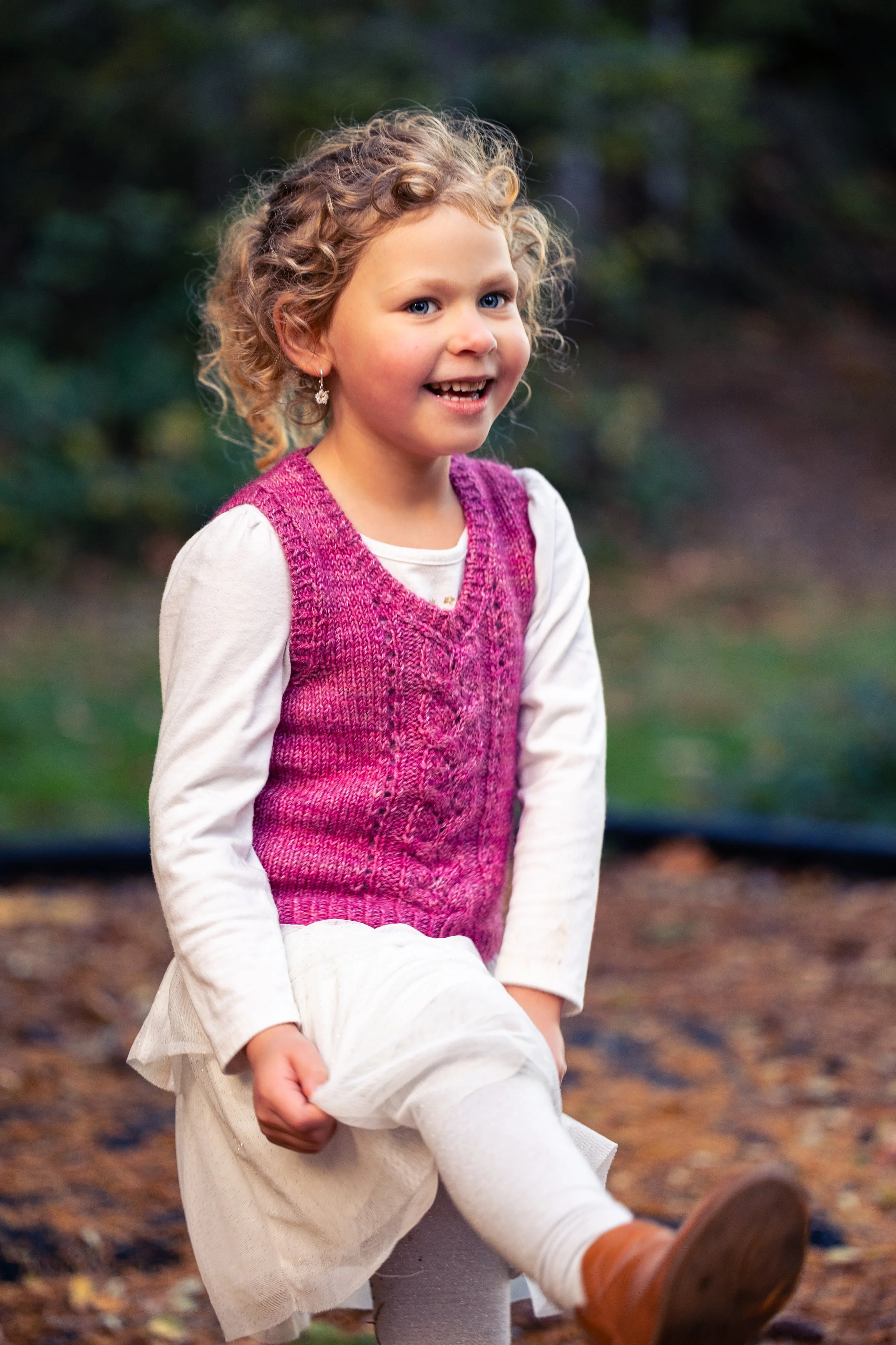 A young child wears a cozy, pink Bittie Bestie Vestie made with a knitting pattern for a kid's V-neck vest by Vanessa Smith Designs. The front features an easy Japanese cable framed by columns of lace