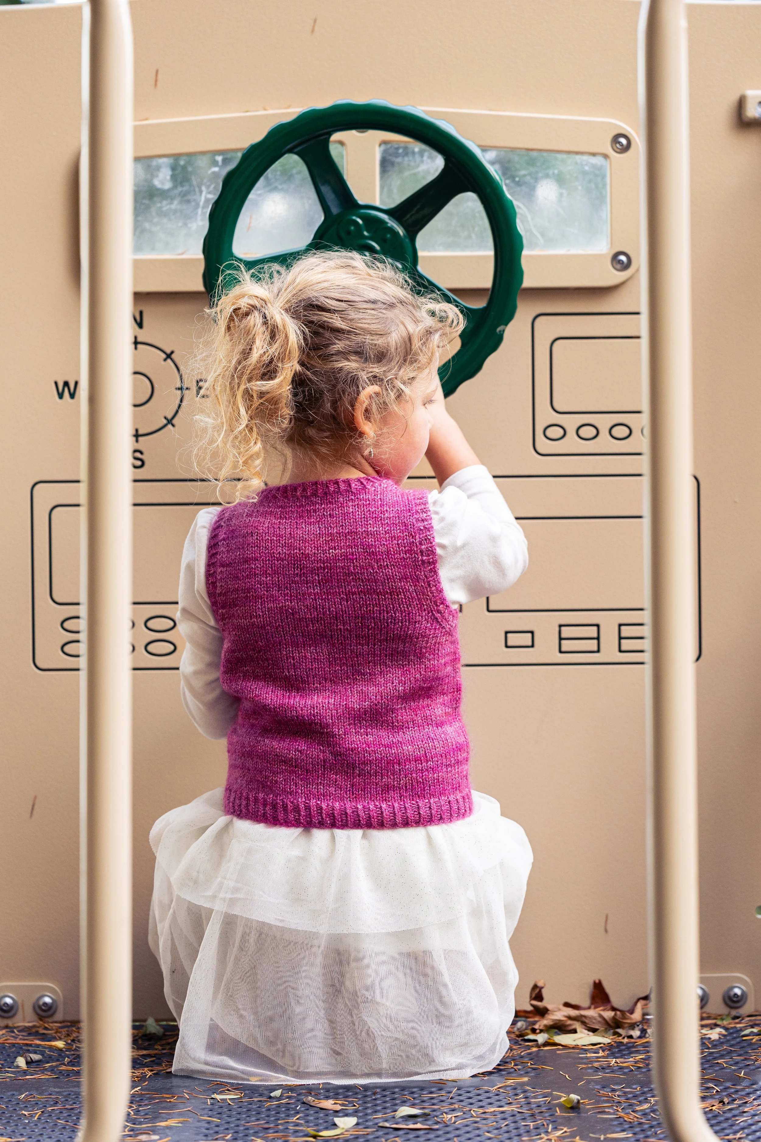 An adorable back view of a rosy pink Bittie Bestie Vestie, sweetly worn over a ruffled dress. Worked from the hem up, this knitting pattern by Vanessa Smith features sizes for babies, toddlers & kids.