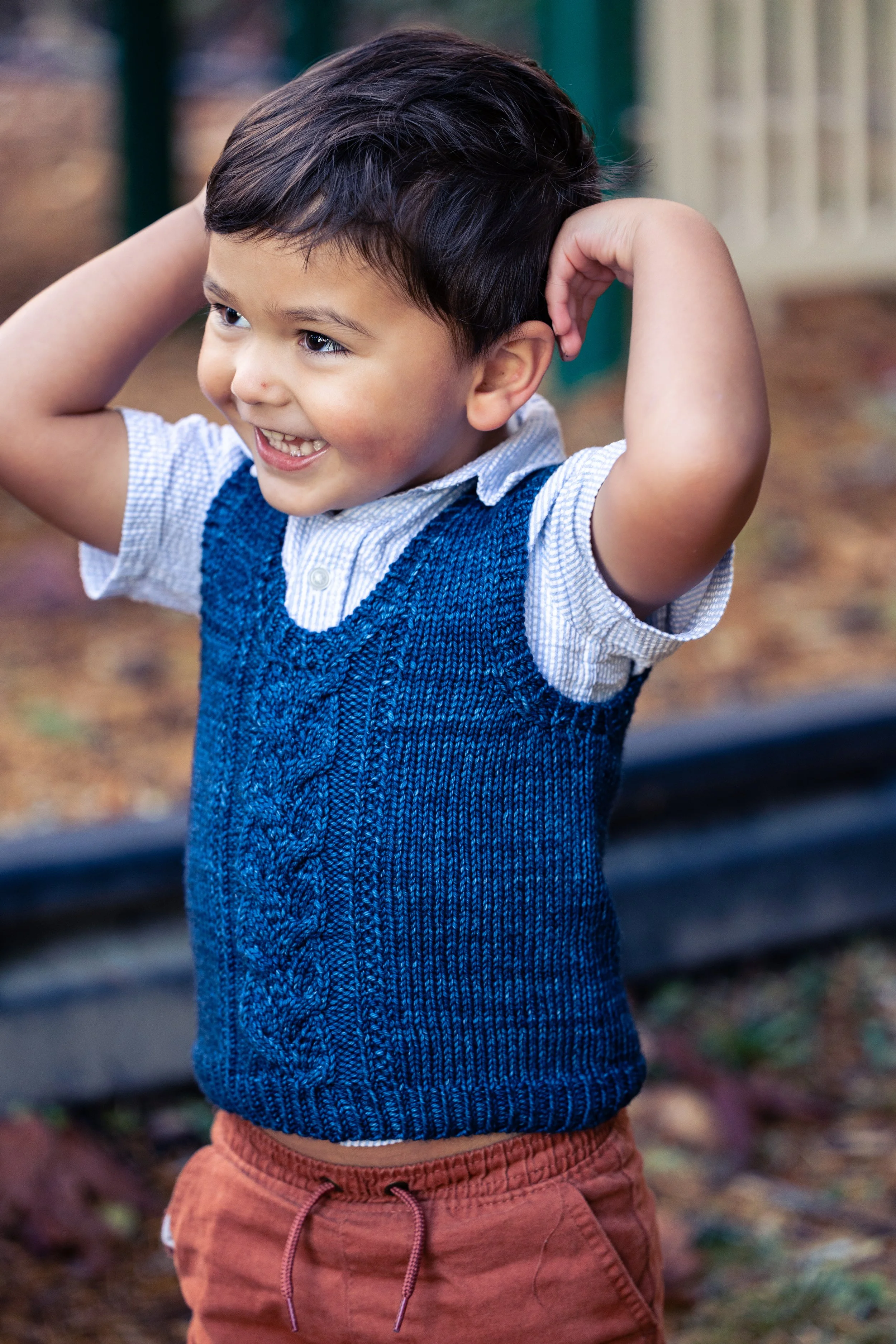 An adorable little boy wears a deep ocean blue, handknit Bittie Bestie Vestie featuring an easy yet striking panel of cables and soft texture. Twisted rib edgings trim the set-in armholes & V neckline