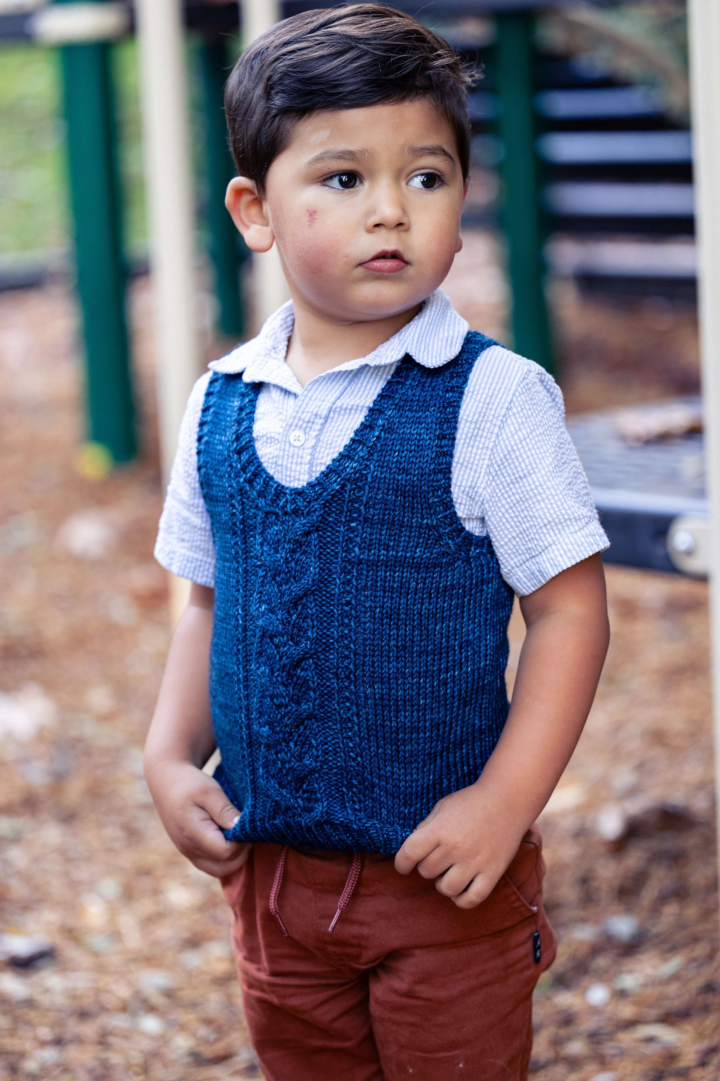 A styled view of the Bittie Bestie Vestie, a classic knit, V-neck sweater vest designed as the perfect layering piece for littles ones. Fun & fast handknit gift idea for baby showers or special events