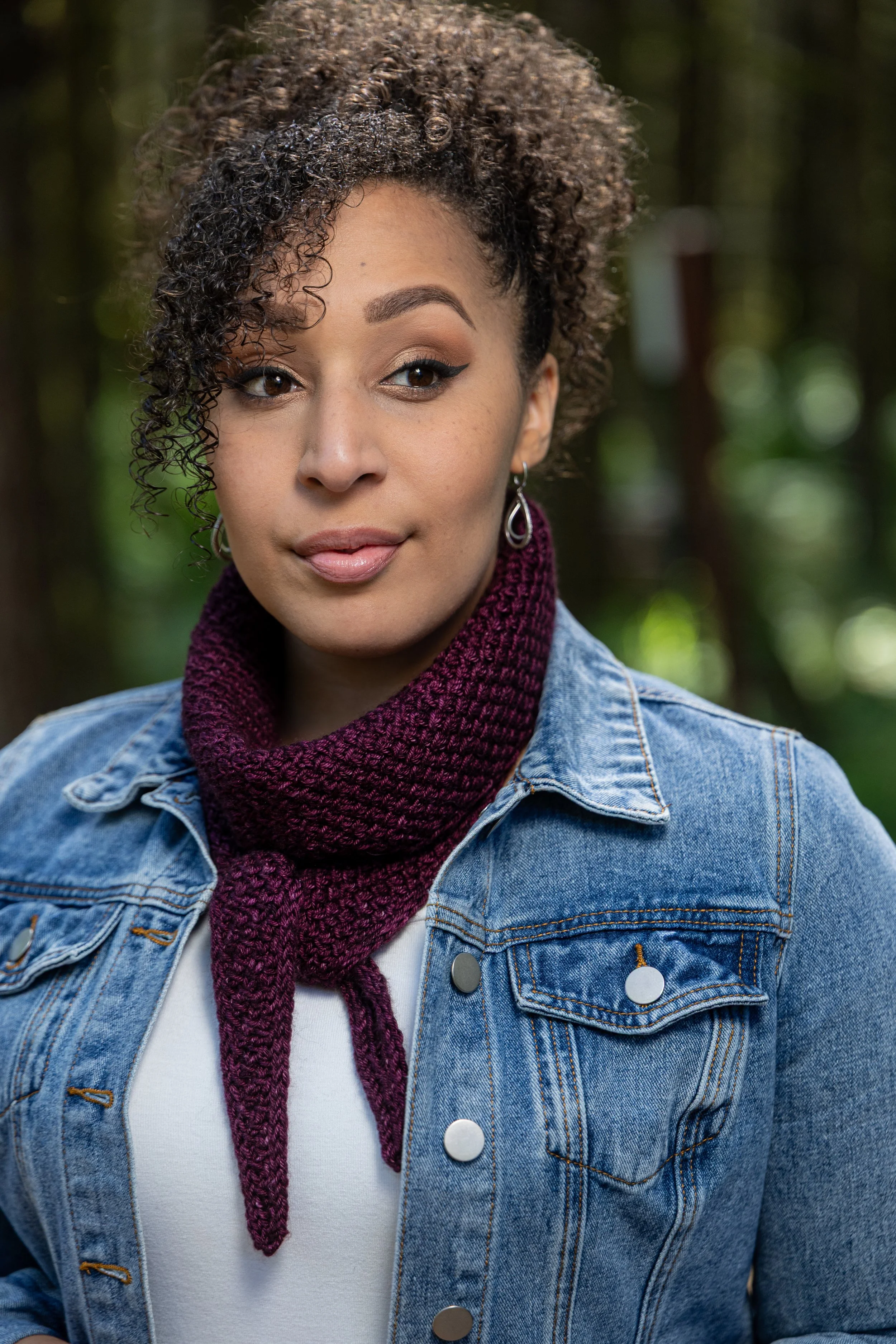 A wine-colored, midi size Tipping Point Scarf worn stylishly knotted as a neck warmer. Features an all-over textured stitch pattern using yarn overs & slipped stitches for a fun, squishy knit fabric.
