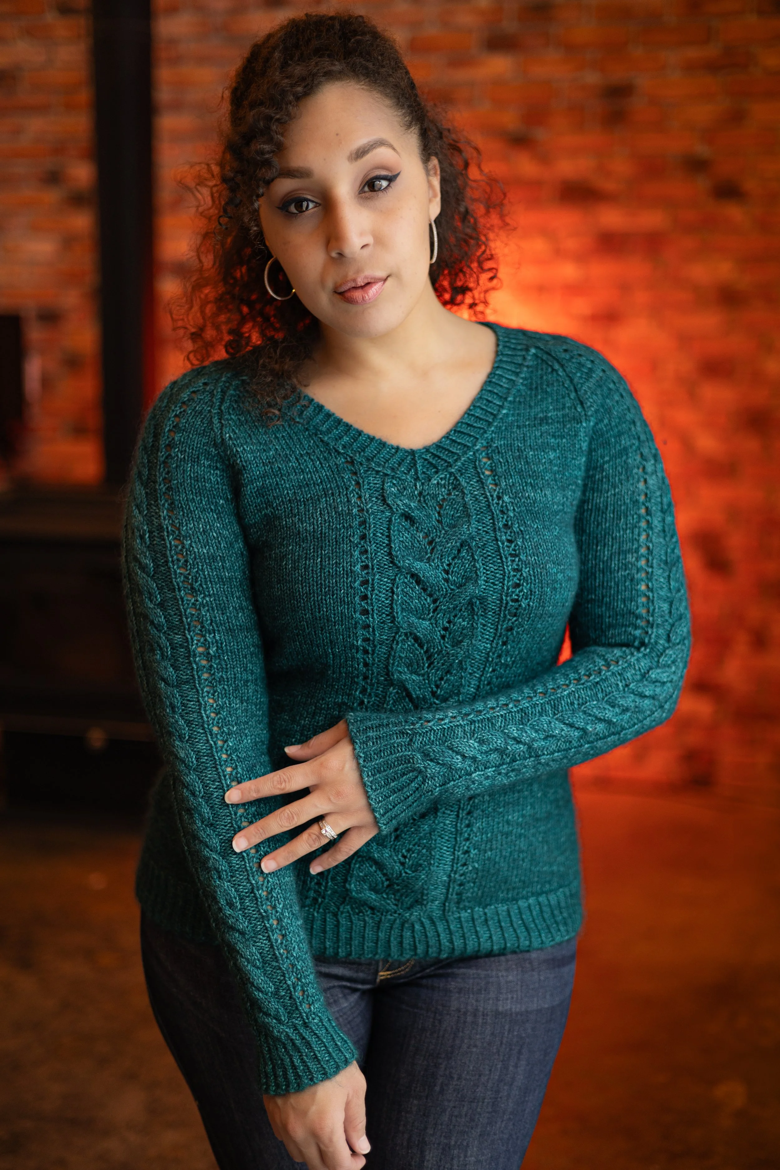 Woman wearing the elegant Meant to Vee sweater in teal, showcasing its V-neck, intricate cable and lace details on sleeves and body. PDF knitting pattern by Vanessa Smith Designs.