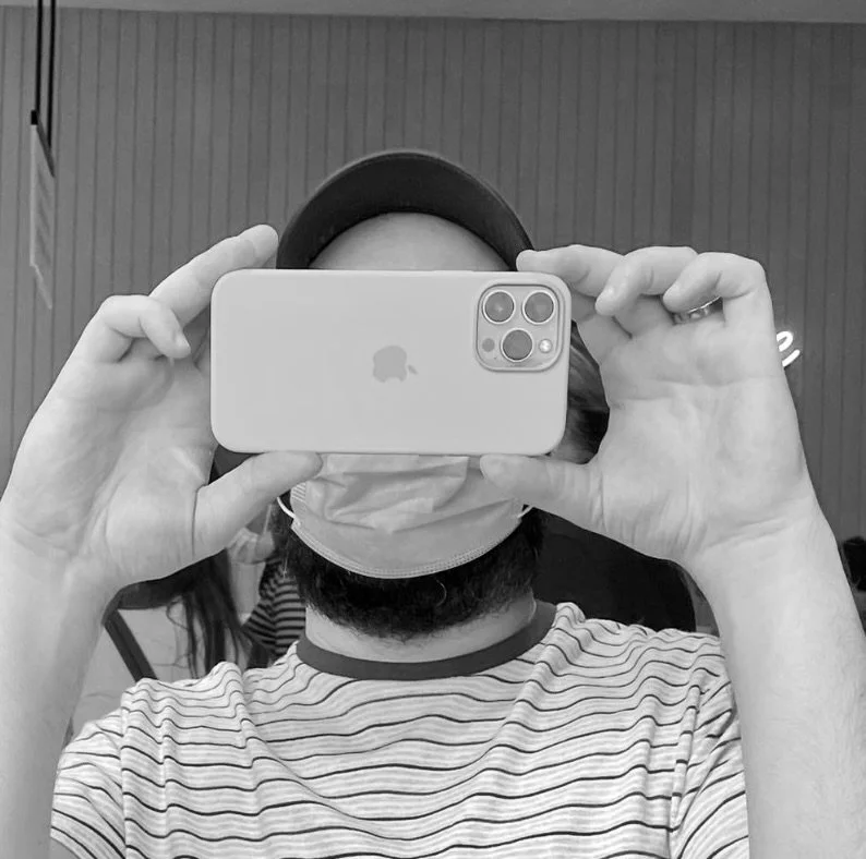 Person taking a mirror selfie with a smartphone, wearing a striped t-shirt, a face mask, and a cap, with a plain background.