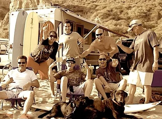 Group of six friends at the beach with two dogs, sitting and standing in front of an RV, enjoying a sunny day.