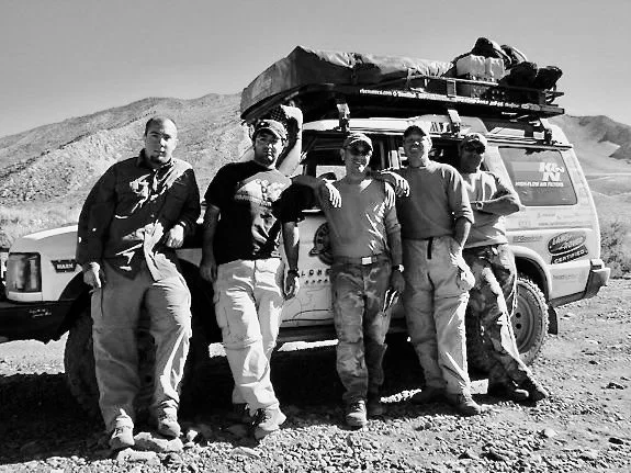 Six young men standing in front of a vehicle with a roof rack and supplies on top, in a desert landscape.