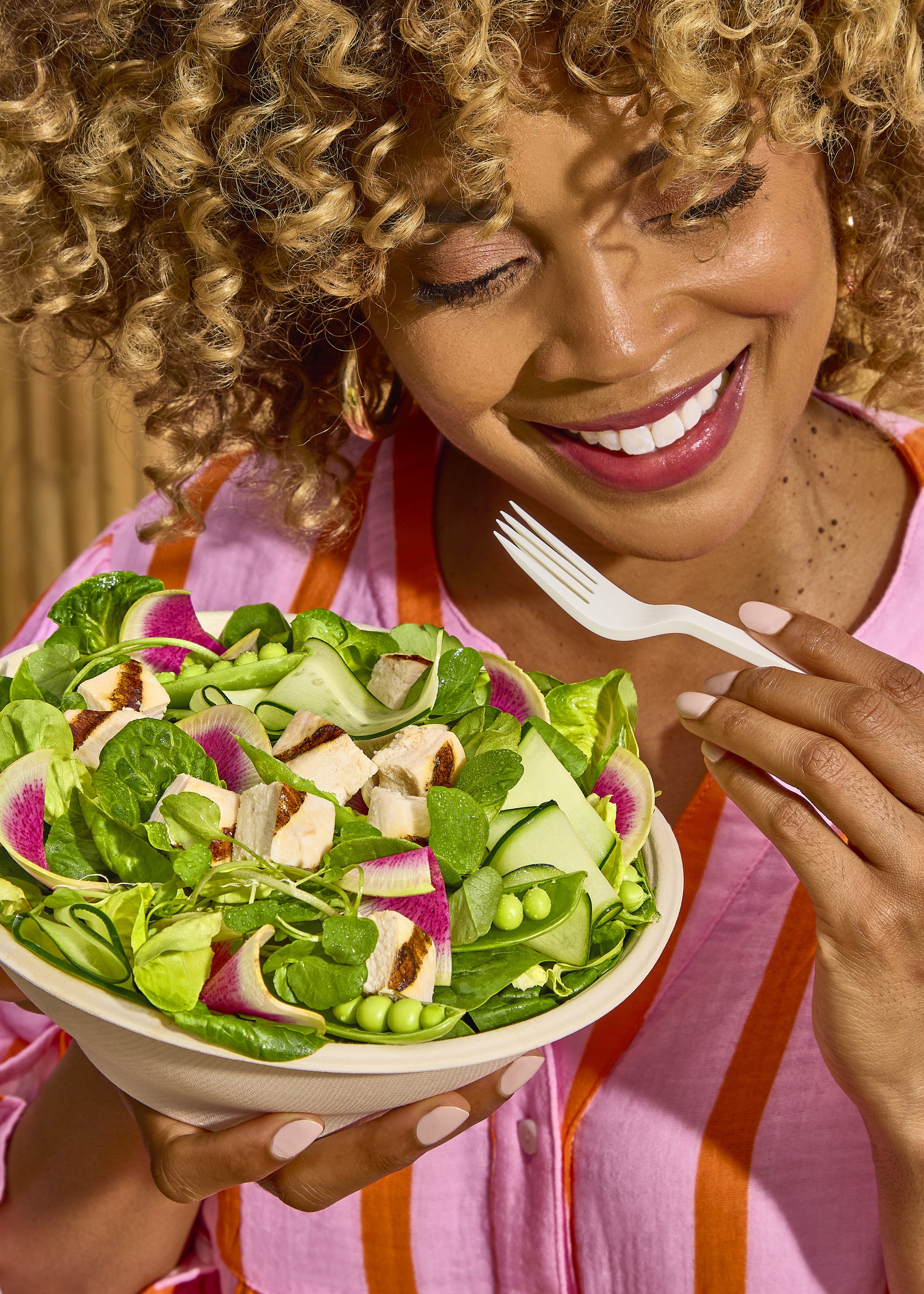 A woman with curly blonde hair smiling and holding a bowl of salad.