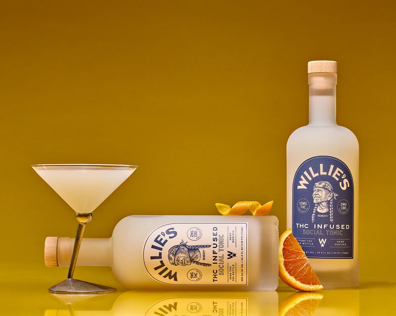 Two bottles of Willie's THC-infused social tonic with a martini glass, orange slices, and a bright yellow background.
