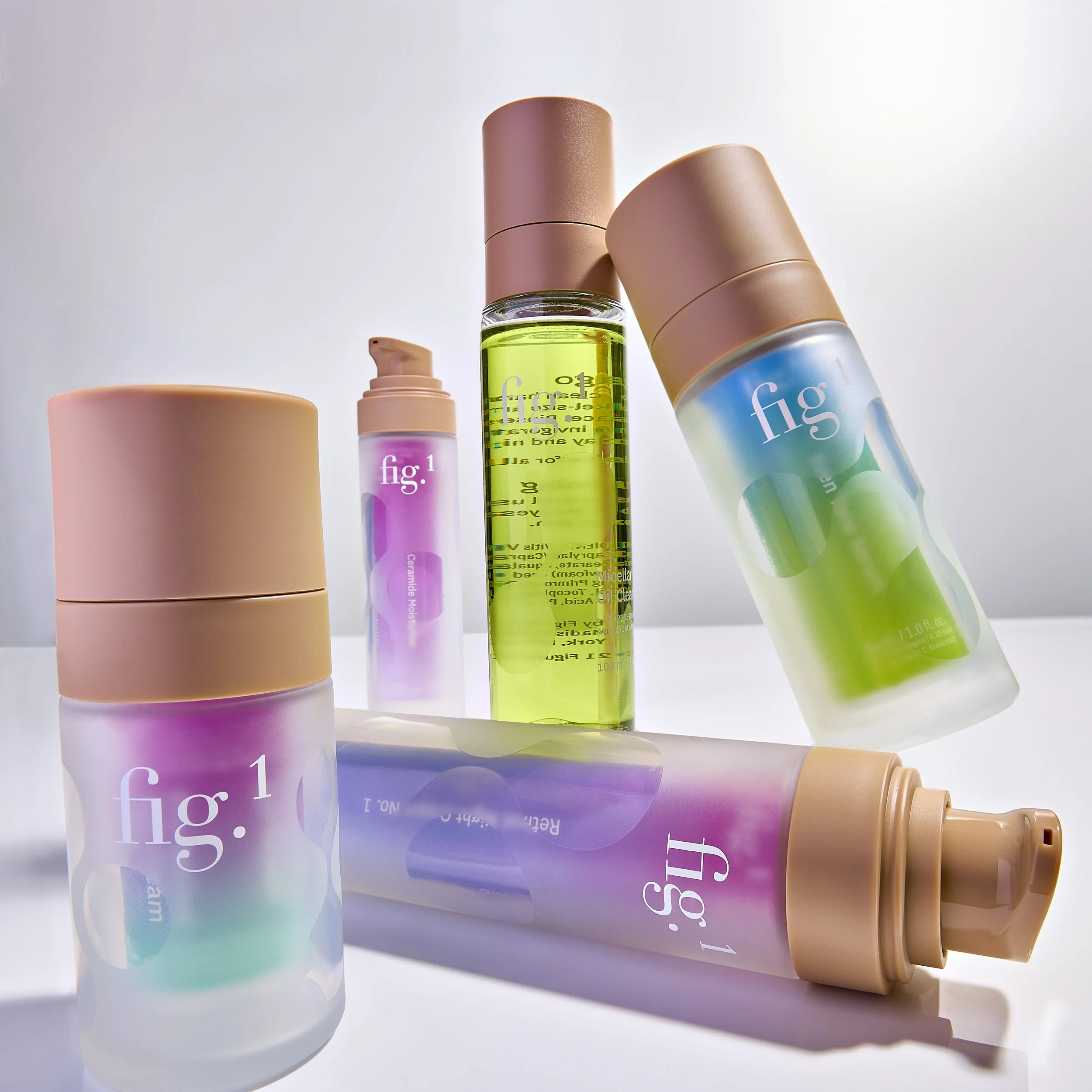 A collection of skincare or cosmetic bottles with pastel and gradient colors, including a pink tube, a pink pump bottle, a green liquid bottle, and a spray bottle, arranged on a white surface.