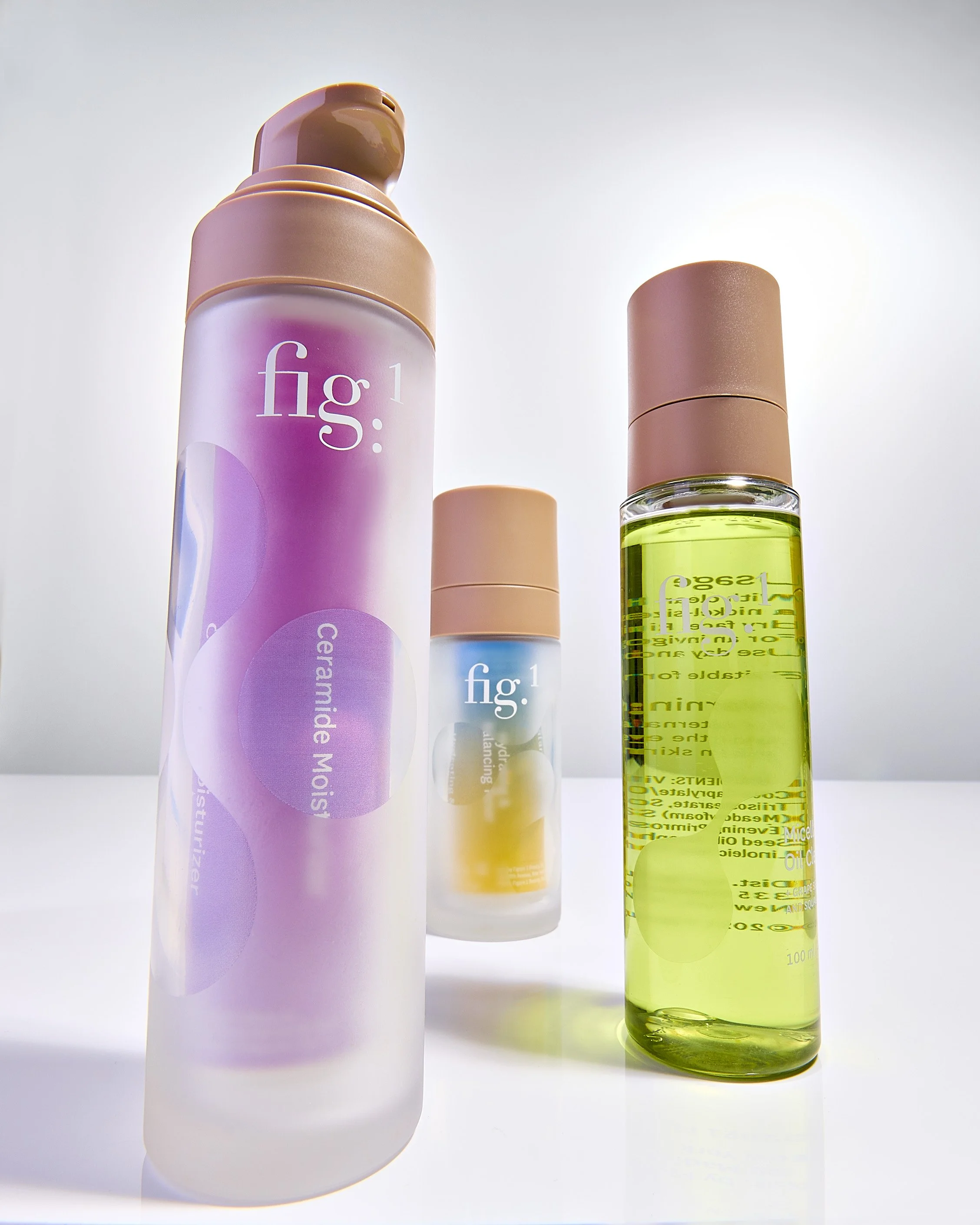 Three cosmetic bottles, the tallest is pink with a beige cap, labeled 'fig1: Ceramide Moisturizer'; the middle is smaller, beige with a beige cap, labeled 'fig1'; the shortest is transparent with a beige cap, containing yellow liquid, labeled 'fig1: 