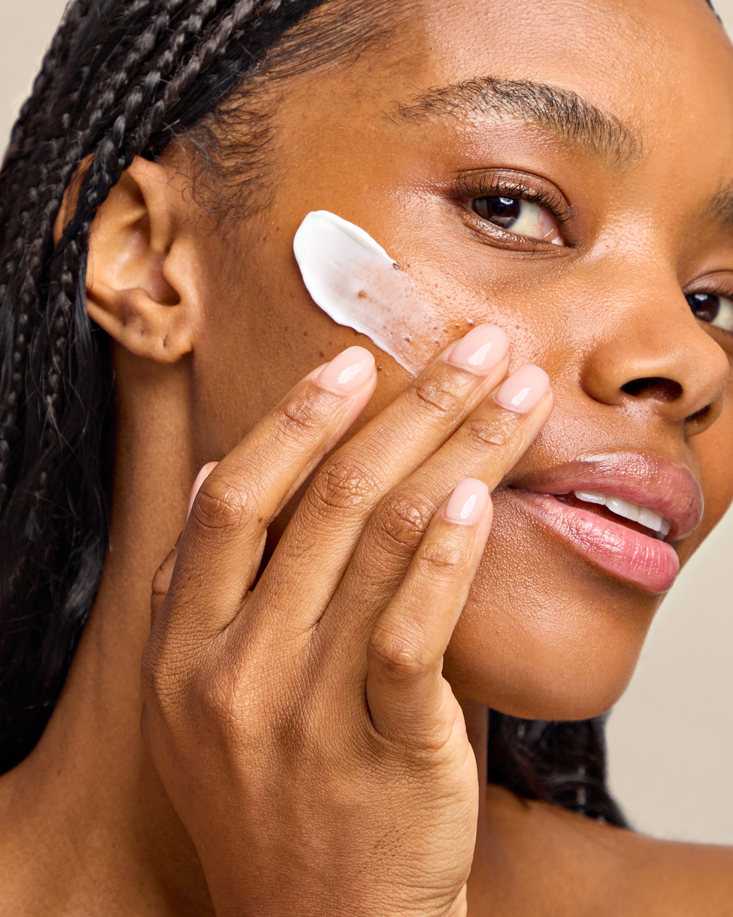 A woman with braided hair applying face cream or moisturizer to her cheek with her fingers.