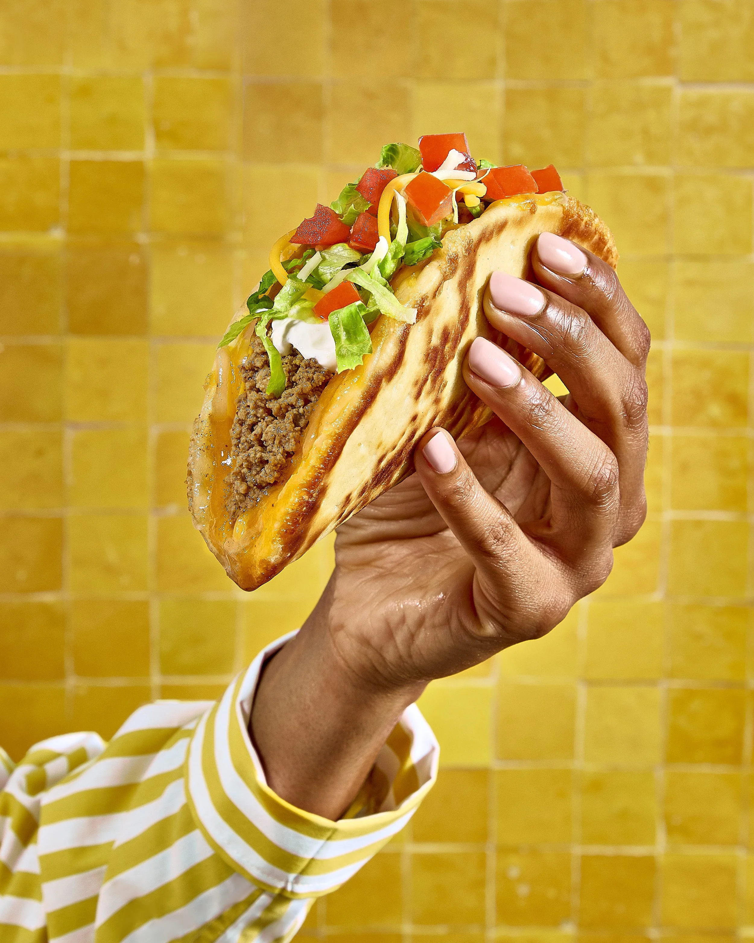 A person with dark skin and pink nails holding a taco with ground beef, shredded cheese, lettuce, diced tomatoes, and sour cream in front of yellow tiled wall.