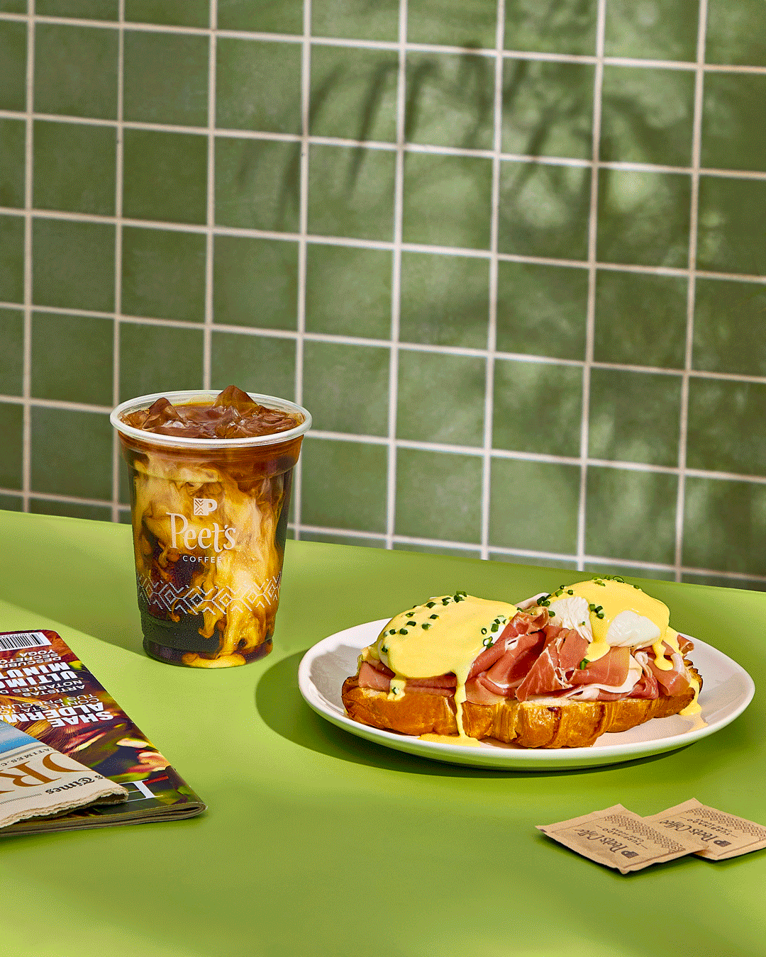 A breakfast setting on a green table with an iced coffee in a Peet's Coffee cup, an open-faced sandwich with eggs, ham, and yellow sauce on toasted bread, and some coffee packets and a magazine.