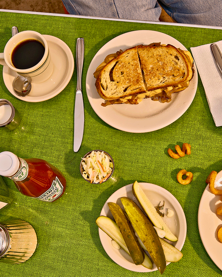 A meal on a green tablecloth includes a cup of black coffee, a plate with a toasted sandwich, a small dish of coleslaw, a plate with pickles and apple slices, and some onion rings with a few scattered on the table.