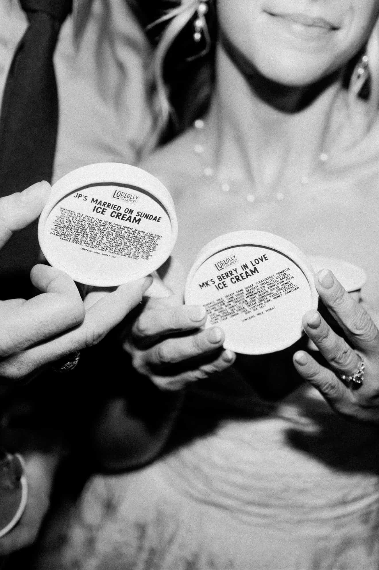 Close-up of bride and groom hands holding ice cream cup lids with custom flavor names, JP's Married on Sundae Ice Cream",  "MK's Berry in Love Ice Cream"