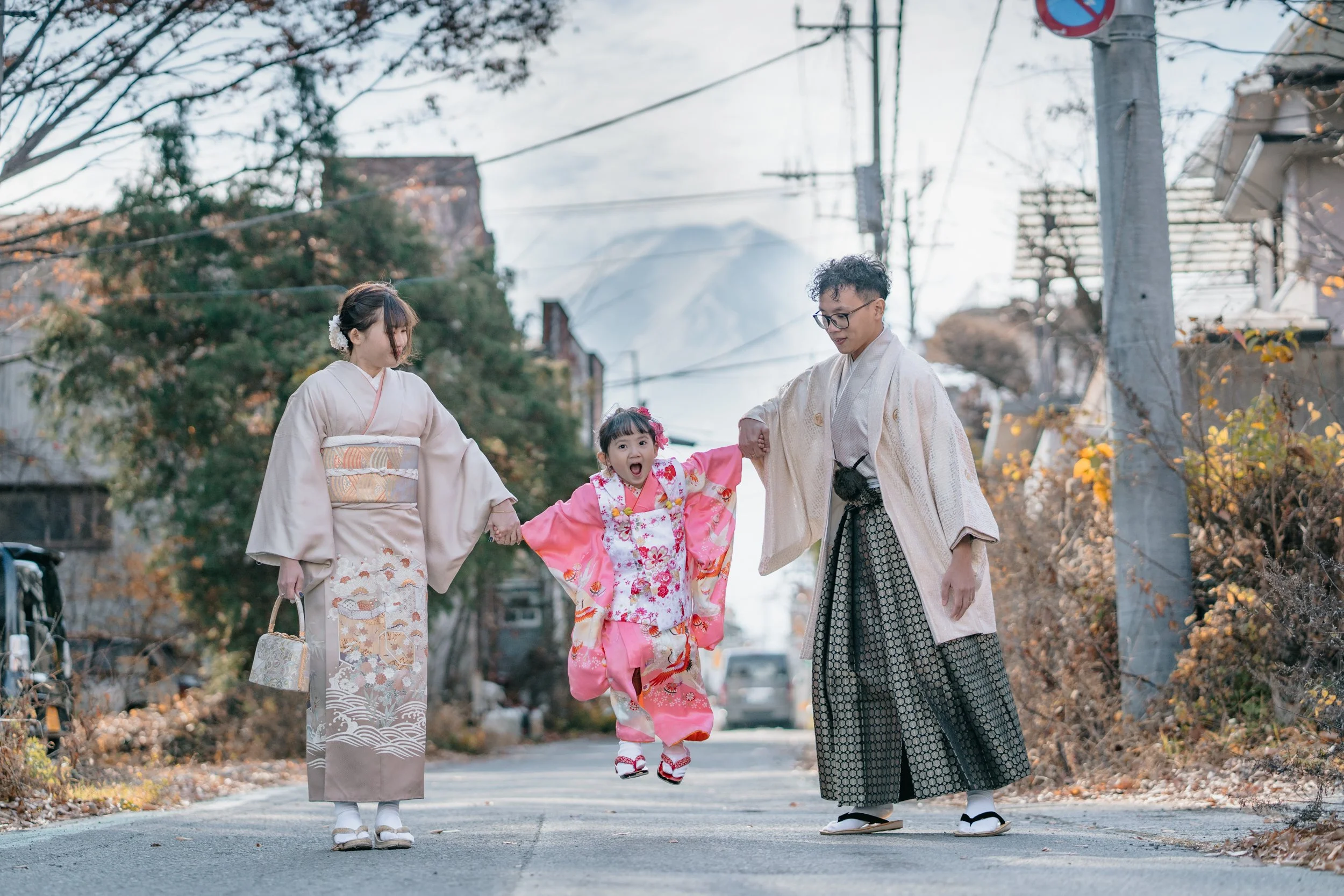 A family of four dressed in traditional Japanese kimono walking together on a street decorated for autumn, with trees and electrical poles in the background.