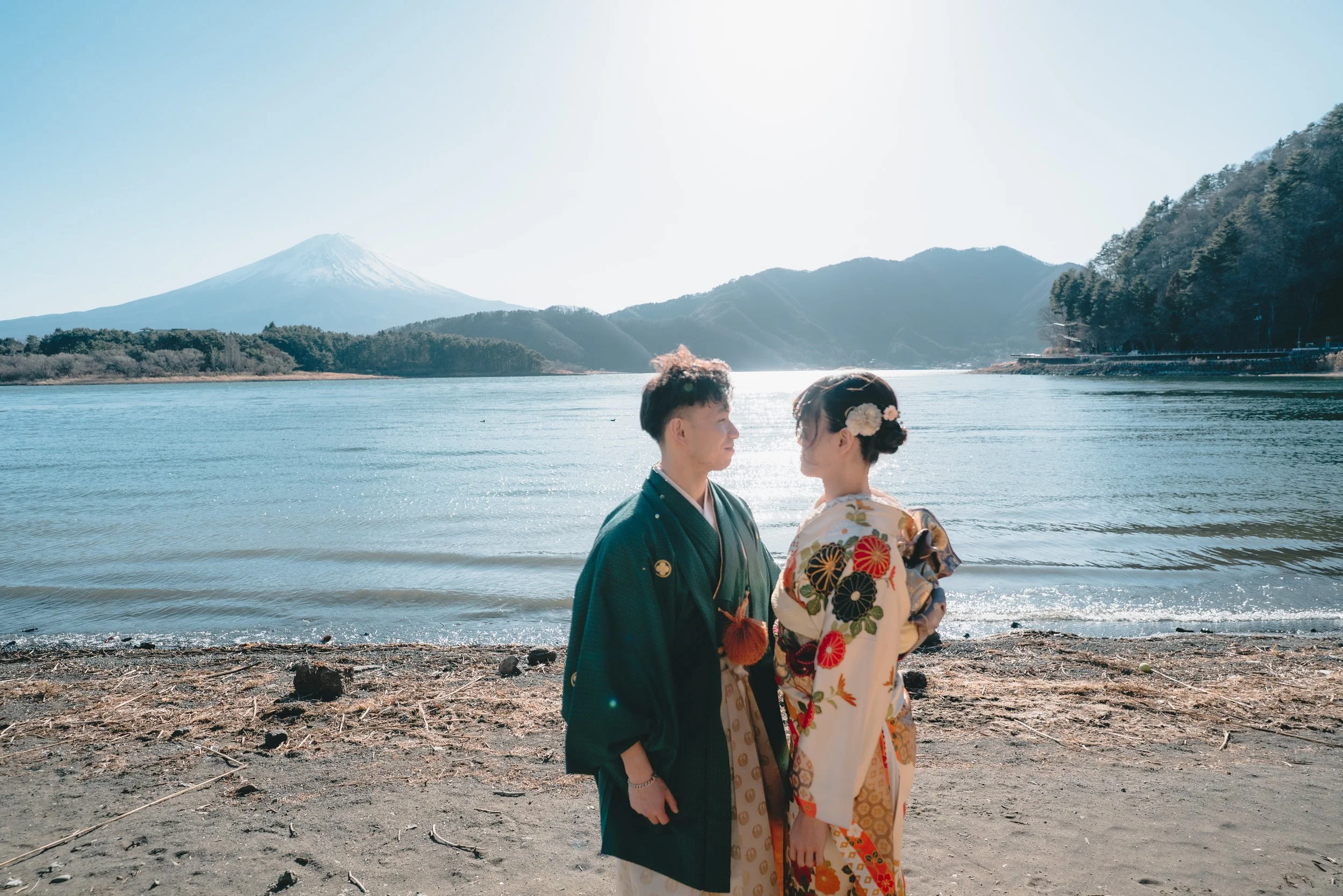 A couple in traditional Japanese attire standing on the shore of a lake with mountains and a snow-capped peak in the background, facing each other.