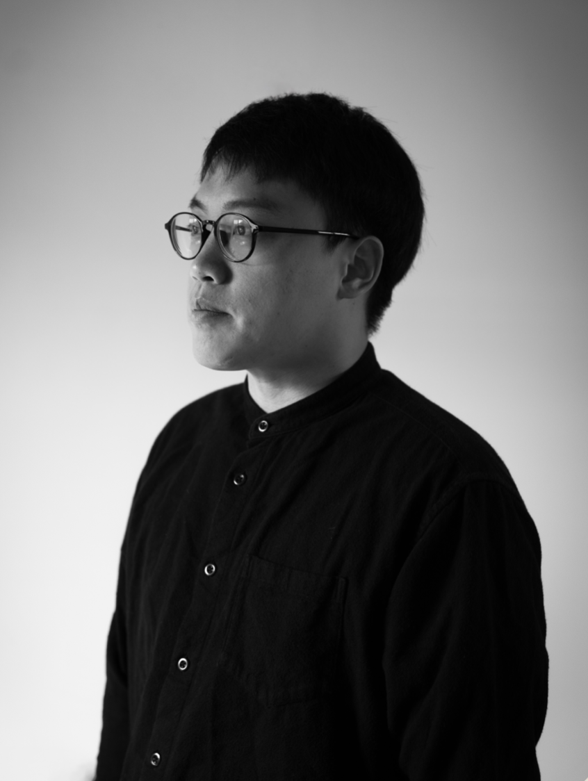 A black and white portrait of a young man with short dark hair and glasses, wearing a dark button-up shirt, facing slightly to the left against a plain light background.